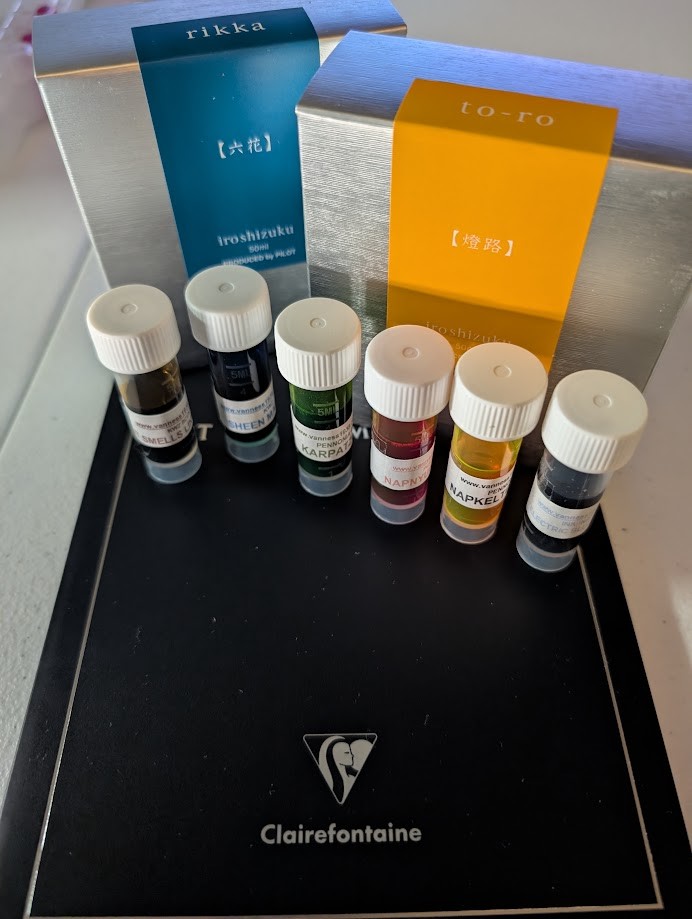 Photo of items on a table: Box with 50ml bottle of Pilot Iroshizuku Rikka fountain pen ink, which is a shade of blue. Box with 50ml bottle of Pilot Iroshizuku To-ro fountain pen ink, which is a shade of yellow-orange. Six sample vials with white caps and labels, the names are not legible but are listed in the main post. A Clairefontaine Triomphe notebook