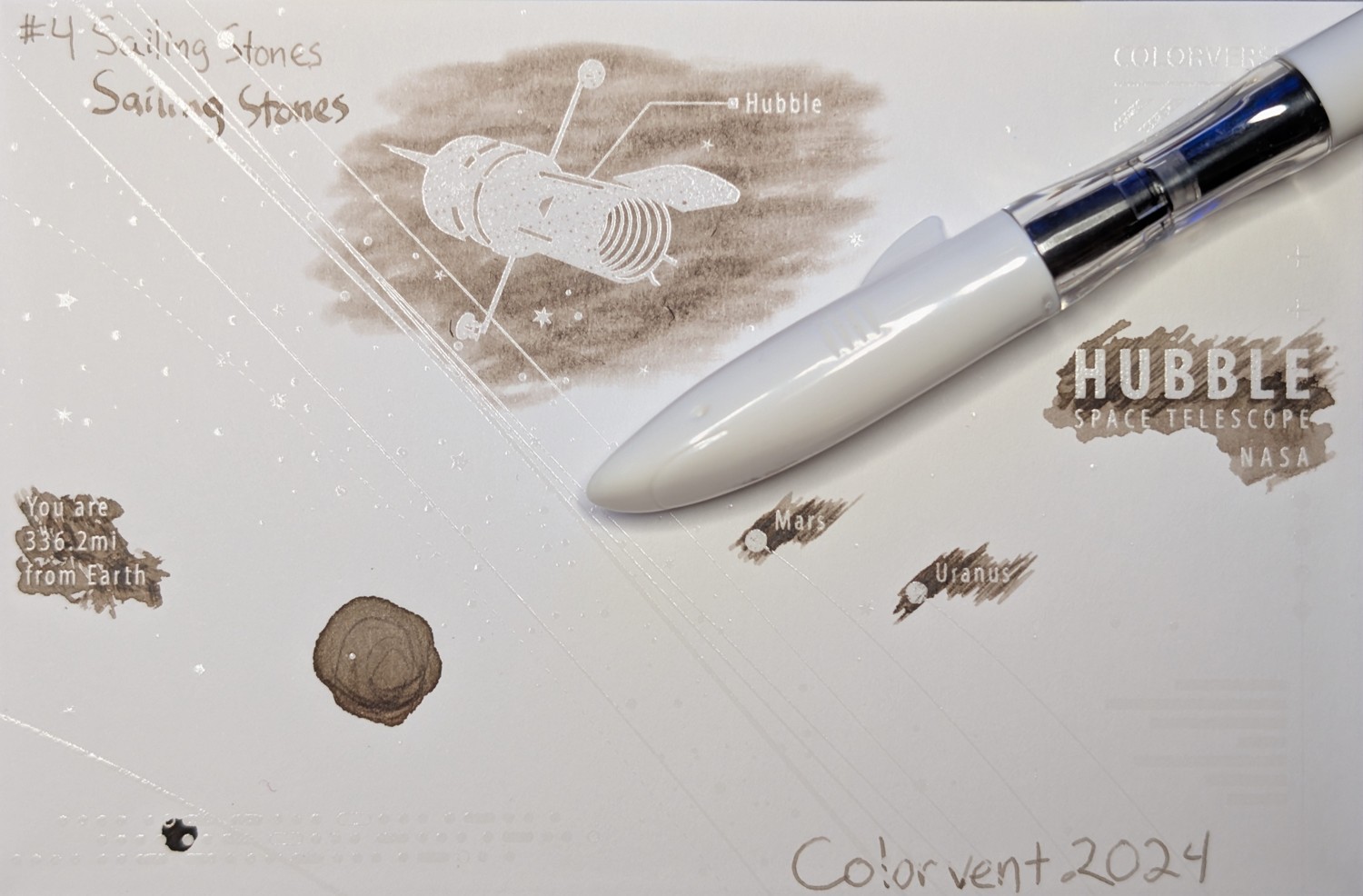 Photo of a "Colorverse Colorspace Size C" swatch card, type 2 which is based on the Hubble space telescope. The ink on the card is a light brown/tan color, with some areas darker where ink has pooled. Upper left is text reading "#4 Sailing Stones" written in an EF nib and then a stub nib. Just to the right side of the middle at the top is a large illustration of the Hubble space telescope in negative space among swabbed ink. In the middle right side is text "Hubble Space Telescope NASA" in negative space among swabbed ink. In the middle left is text reading "You are 336.2mi from Earth" in negative space among ink scribbles. In the middle lower left is a blob of swirled ink drawn by a glass dip pen. Near the center are "Mars" and "Uranus" with circles representing those planets, in more ink. Lower right is text "Colorvent 2024". Laying across the card is a white Jinhao Shark fountain pen with a clear section. The cap of the pen is shaped like the front of a shark, with a mouth, eyes, and a fin.