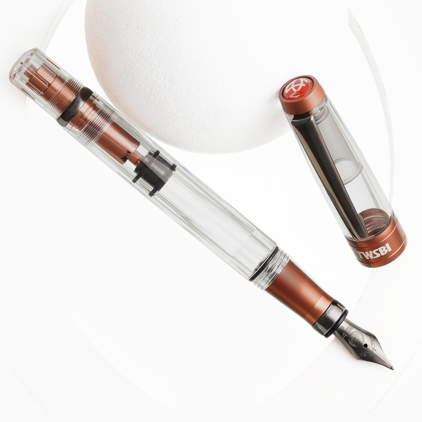Photo of a fountain pen next to a circular bit of foam(?) that may be a partial sphere. The cap is off and resting on the foam. The pen has a clear body with brown metal about the same color as coffee around the grip section and inside the piston knob, with dark "onyx" trim and nib.