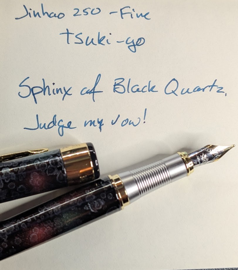 Photo of a writing sample and fountain pen. The pen is uncapped and the cap is laying next to the pen. The writing sample is in ink that is dark blue with a hint of green. It reads "Jinhao 250 - Fine / Tsuki-yo / Sphinx of Black Quartz, Judge my vow!". The pen is a dark base color, blue nearly black. There are curcular bursts of red and green on the pen. There are also whiteish areas that look like various shapes of droplets with thicker outlines. The section on the pen is silver and ribbed. The nib is two-tone silver and gold plating. The accents on the cap and around the section are gold.
