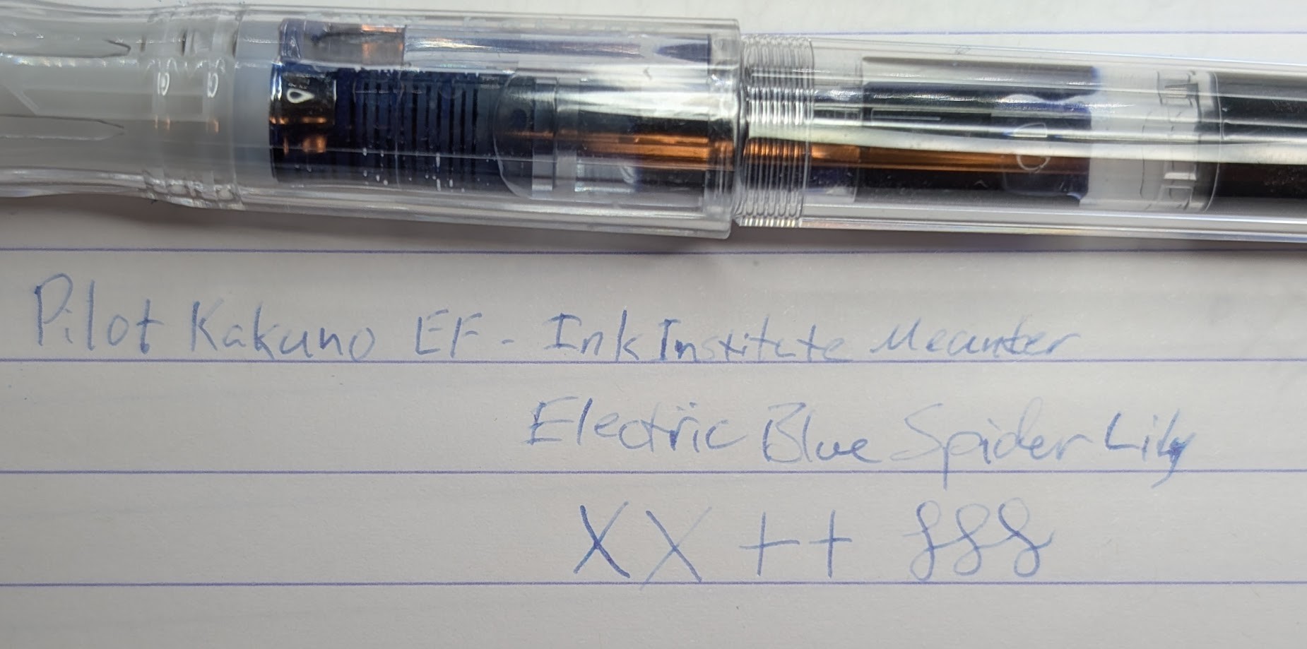 Photo of a clear Pilot Kakuno fountain pen above a writing sample on a piece of lined paper. The writing sample is in very faint light blue ink with extremely thin line width reading "Pilot Kakuno EF - Ink Institute Meander / Electric Blue Spider Lily" followed by "XX ++" and a few curlicues.
