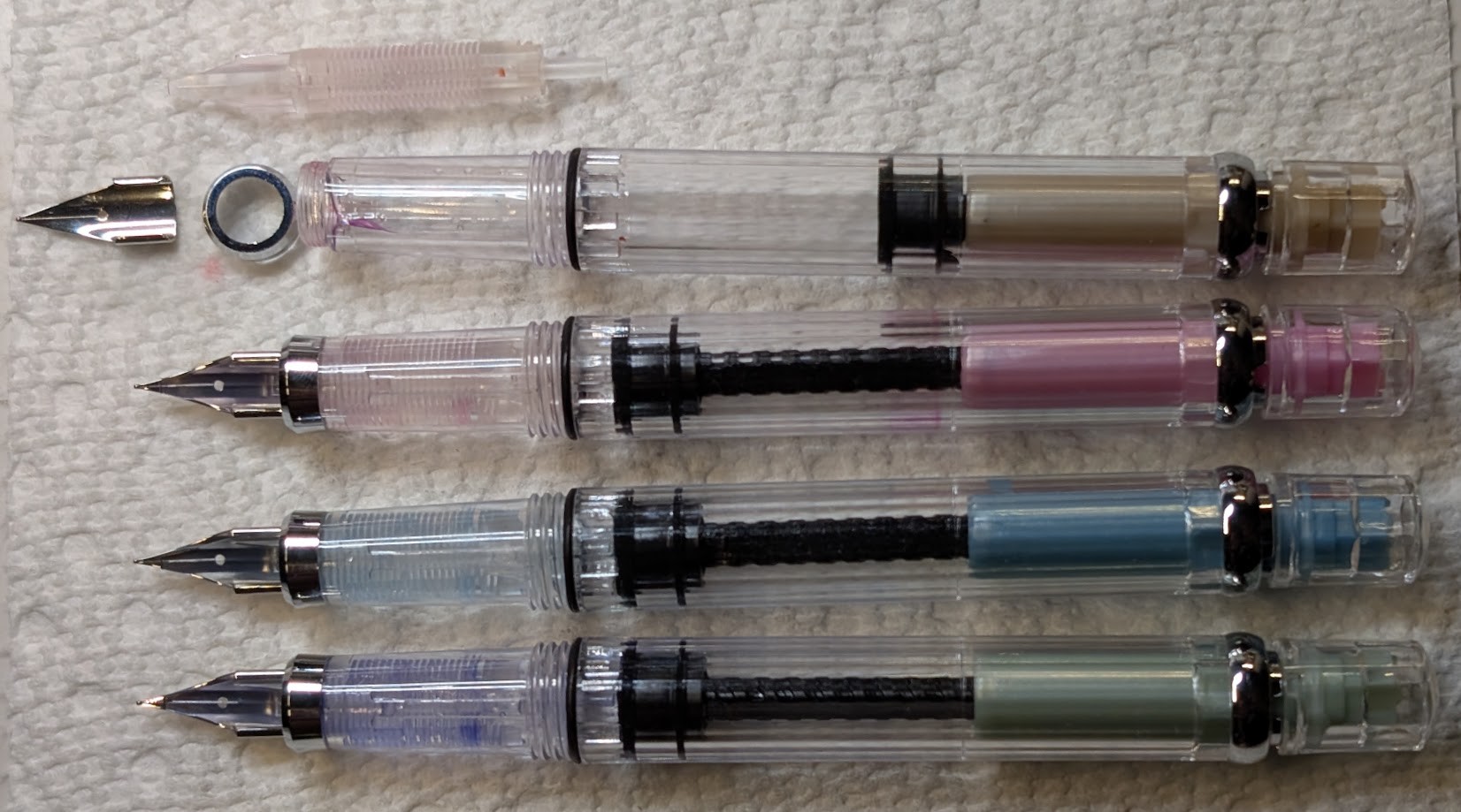Four uncapped piston filler fountain pens in a row. The top pen has orange accents and has the nib, feed, and a collar removed. There is a visible crack near the tip. Second pen has pink accents and slight red staining in the feed. Third pen has light blue accents and light blue staining in the feed. Last pen has green-ish(?) accents and blurple staining in the feed.