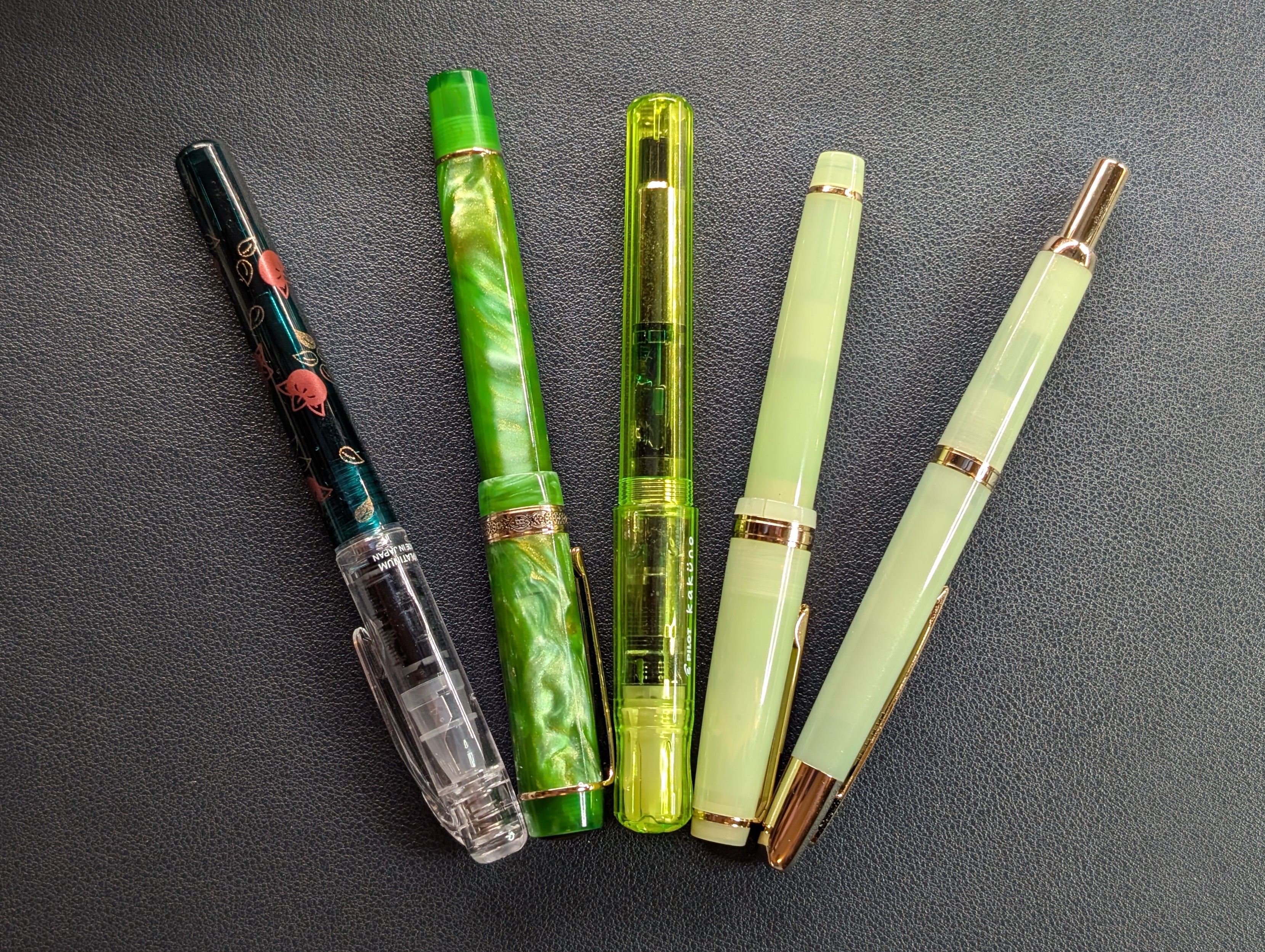Photo of five fountain pens arranged with their tips touching and radiating away from the bottom center. From left to right the pens are: - A dark green Platinum Preppy with a printed design (Preppy Wa Modern Maki-e Tachibana) - A bright green Kaigelu 316A with a shimmery swirl that has varying tints and shades of green (Dream Green Swirl Marble) - A transparent yellow-green Pilot Kakuno (Kakuno Family Series Baby Yellow) - A semi-transparent pale light green Jinhao 82 (Clear Tender Green), there is a slight crack at the base of its cap - A semi-transparent pale light green Jinhao 20 (Celestial Jade)