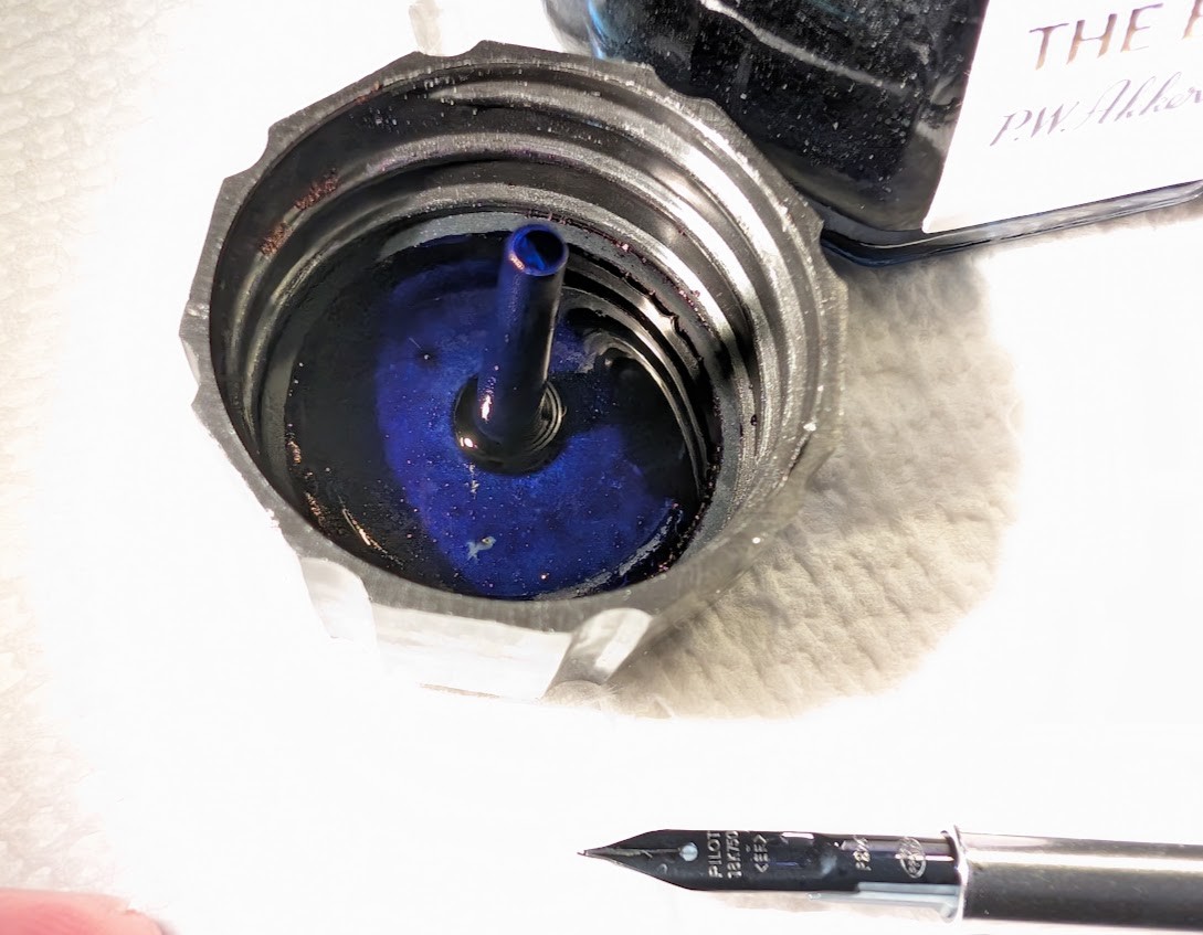 Overexposed photo of a fountain pen ink bottle cap, inverted and laying on a paper towel next to a Pilot VP fountain pen nib unit. The cap has a long cylinder rising up from the underside of the lid, extending well past the height of the threaded part of the cap. The underside of the ink and that cylinder are coated in dark blue/purple ink.