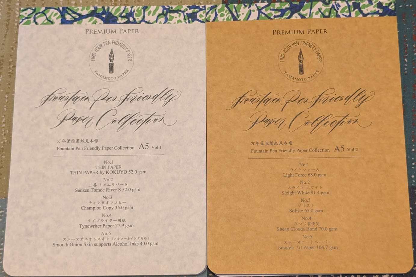 Photo of two pads of paper showing their covers. They are of similar design. The left pad has a light marble-like finish and has the following text: Premium Paper, Find your pen friendly paper, Yamamoto Paper. Fountain Pen Friendly Paper Collection A5 Volume 1. No 1. Thin Paper by Kokuyo, 52.0 gsm. No 2. Sanzen Tomoe River S 52.0gsm. No 3. Champion Copy 35.0gsm. No. 4 Typewriter Paper 27.9gsm. No. 5 Smooth Onion Skin supports Alcohol Inks 40.0gsm The right pad has an ivory-colored marble-like finish and has the following text: Premium Paper, Find your pen friendly paper, Yamamoto Paper. Fountain Pen Friendly Paper Collection A5 Volume 2. No 1. Light Force 68.0gsm. No. 2 Sleight White 81.4gsm. No 3. Soliste 65.0gsm. No. 4 Sheep Clouds Bond 70.0gsm. No. 5 Smooth Art Paper 104.7gsm.