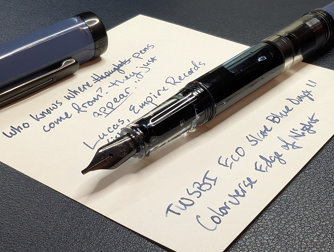 Photo of a desk containing a fountain pen and writing sample on a piece of note paper. The photo is taken from the front left corner at a shallow angle and is and focused on the pen nib. The fountain pen is uncapped, the cap a dark blue-gray color and is laying across the top of the paper. The pen has a black nib, clear section and body, and a dark blue-gray tail. It is a piston filling pen with blue ink visible inside. The TWSBI name and logo are visible, engraved into the nib. The writing sample reads: "Who knows where thoughts come from? They just appear." Thoughts is crossed out and replaced by "pens". The quote is attributed to "Lucas (ish), Empire Records" There is more writing under the pen: "TWSBI ECO Slate Blue Onyx 1.1", "Colorverse Edge of Night"