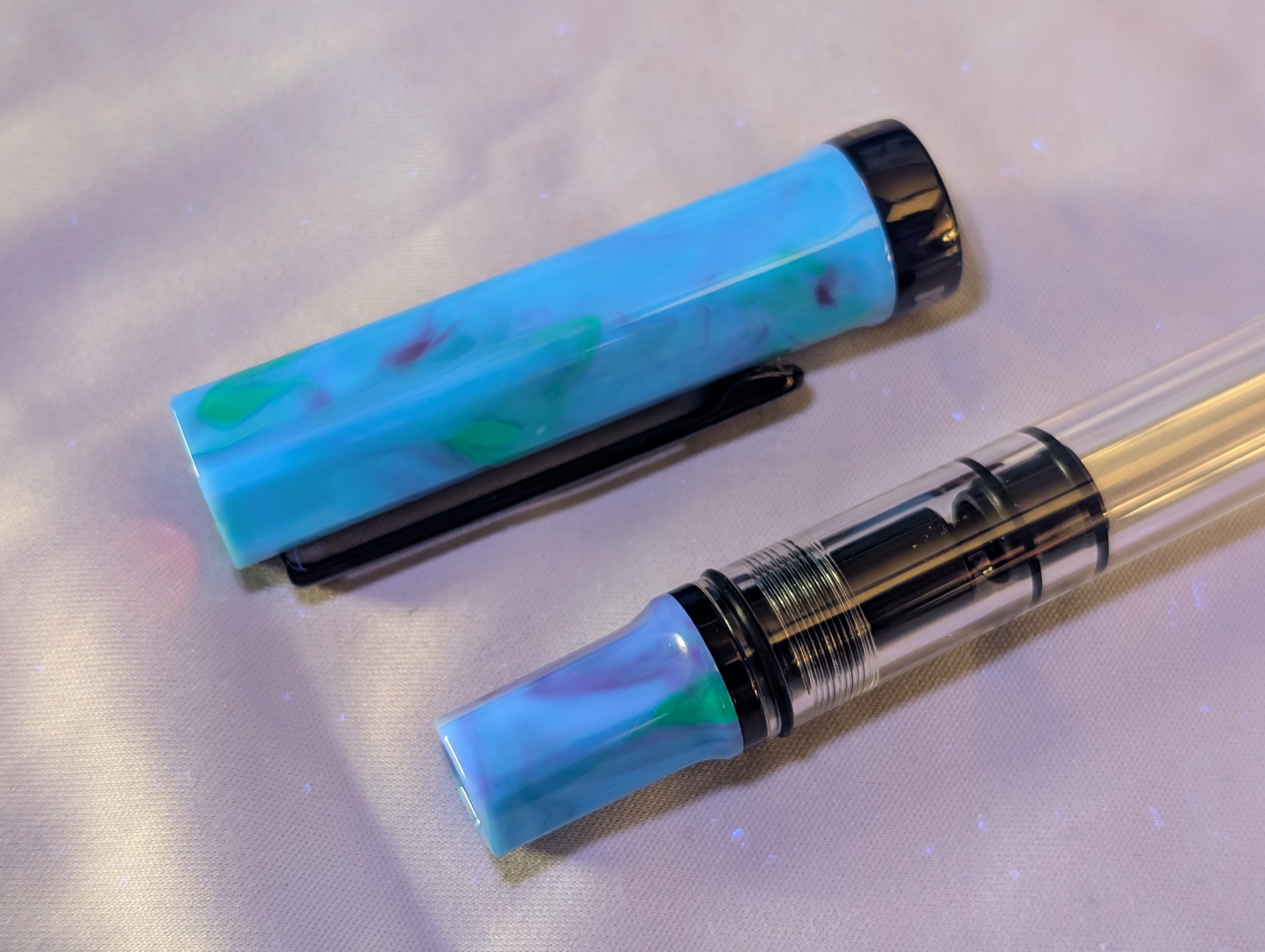 Photo under UV light of an uncapped fountain pen with the cap laying next to the pen. The tail end of the pen is situated next to the cap. The pen cap and piston knob are a swirled mix of blue with streaks of purple and green, which is glowing and brighter than the previous photo.