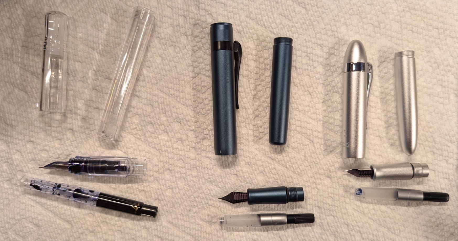 Photo of three disassembled fountain pens on a paper towel. A Pilot Kakuno with a CON-70 conveter, a blue Hongdian M2, and a silver Hongdian M1.
