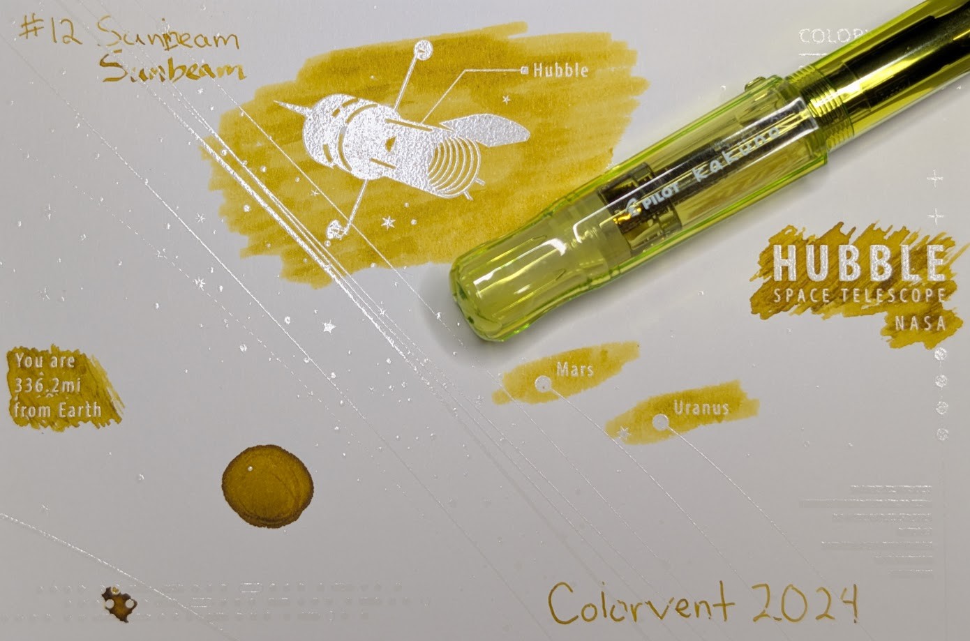 Photo of a "Colorverse Colorspace Size C" swatch card, type 2 which is based on the Hubble space telescope. The ink on the card is a dark yellow-brown with darker shading areas where ink has pooled. Upper left is text reading "#12 Sunbeam" written in an EF nib and then a stub nib. Just to the left side of the middle at the top is a large illustration of the Hubble space telescope in negative space among swabbed ink. In the middle right side is text "Hubble Space Telescope NASA" in negative space among scribbled ink. In the middle left is text reading "You are 336.2mi from Earth" in negative space among ink scribbles. In the middle lower left is a blob of swirled ink drawn by a glass dip pen. Near the center are "Mars" and "Uranus" with circles representing those planets, in more swabbed ink. Lower right is text "Colorvent 2024". Laying across the card is a Pilot Kakuno fountain pen, which is transparent yellow-green.