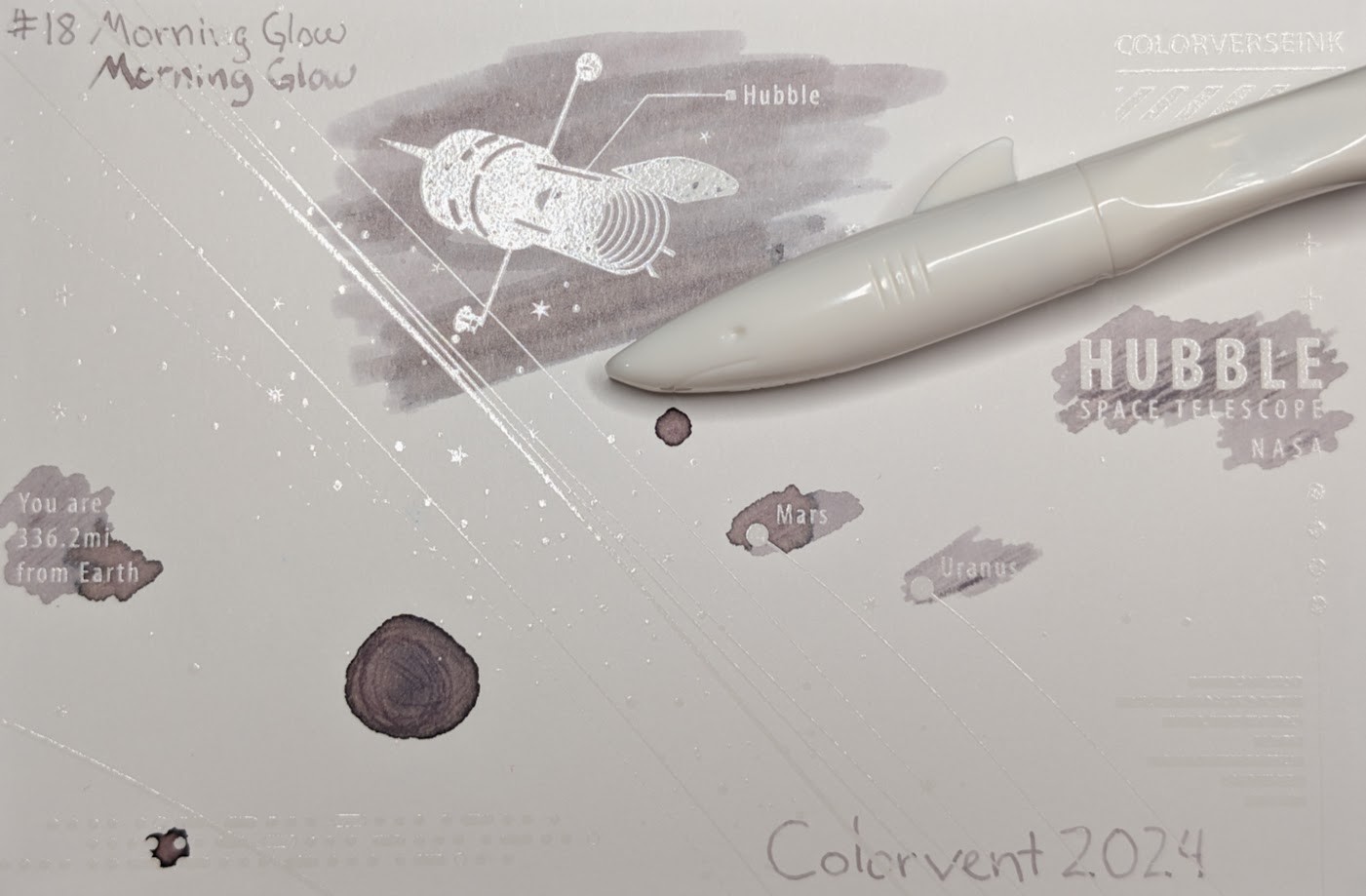 Photo of a "Colorverse Colorspace Size C" swatch card, type 2 which is based on the Hubble space telescope. The ink on the card is a light gray with some darker areas were more ink settled on the paper. Those darker areas vary in color but most have a pinkish or brownish tone. Upper left is text reading "#18 Morning Glow" written in an EF nib and then a stub nib. Just to the left side of the middle at the top is a large illustration of the Hubble space telescope in negative space among swabbed ink. In the middle right side is text "Hubble Space Telescope NASA" in negative space among scribbled ink. In the middle left is text reading "You are 336.2mi from Earth" in negative space among ink scribbles. In the middle lower left is a blob of swirled ink drawn by a glass dip pen. Near the center are "Mars" and "Uranus" with circles representing those planets, in more swabbed ink. Lower right is text "Colorvent 2024". Laying across the card is a white Jinhao Shark fountain pen with a white section. The cap of the pen is shaped like the front of a shark, with a mouth, eyes, and a fin.