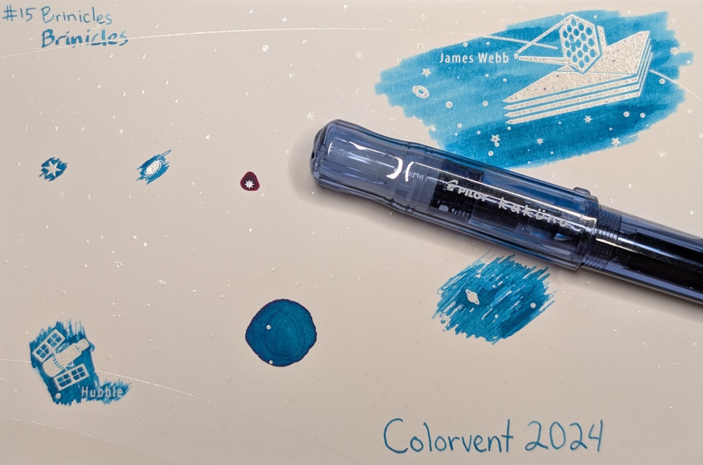 Photo of a "Colorverse Colorspace Size C" swatch card, type 1 which is based on the James Webb space telescope. The ink on the card is medium blue-green that shades a little and has a tiny bit of reddish sheen. Upper left is text reading "#15 Brinicles" written in an EF nib and then a stub nib. Below the text are multiple stars and a galaxy outlined in ink. Upper right is a large illustration of the James Webb telescope in negative space among swabbed ink. In the lower left is a small illustration of the Hubble space telescope in scribbled ink. In the middle lower left is a blob of ink with some visible areas where it dried darker and has reddish sheen. There are lines of darker ink in the center. In the middle right is a blob of scribbled ink drawn by a glass dip pen. Lower right is text "Colorvent 2024". Laying across the card is a fountain pen, a Pilot Kakuno Family Dad (Blue) which is transparent blue