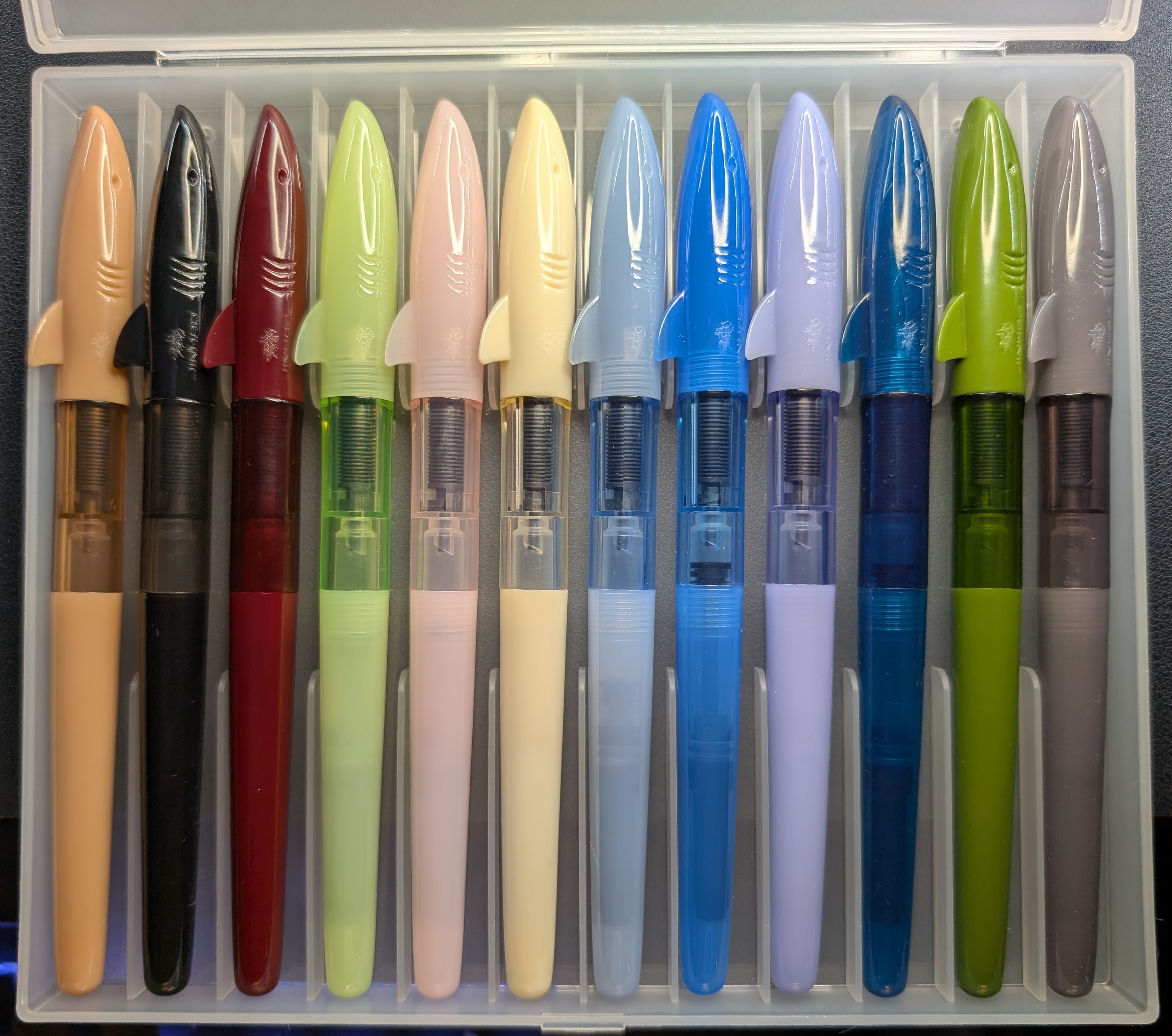 Photo of a case containing 12 Jinhao Shark fountain pens. They have a cap made to look like the front half of a shark (complete with a fin roll stop, gills, eyes, and a mouth). They have long tapered bodies. Each has a tinted but transparent section in the middle, giving a good view of the feed and tip of the converter. From left to right the colors are: tan, black, maroon, mint green, pink, pale yellow, light blue, medium blue, lavender, dark blue, green, and gray.