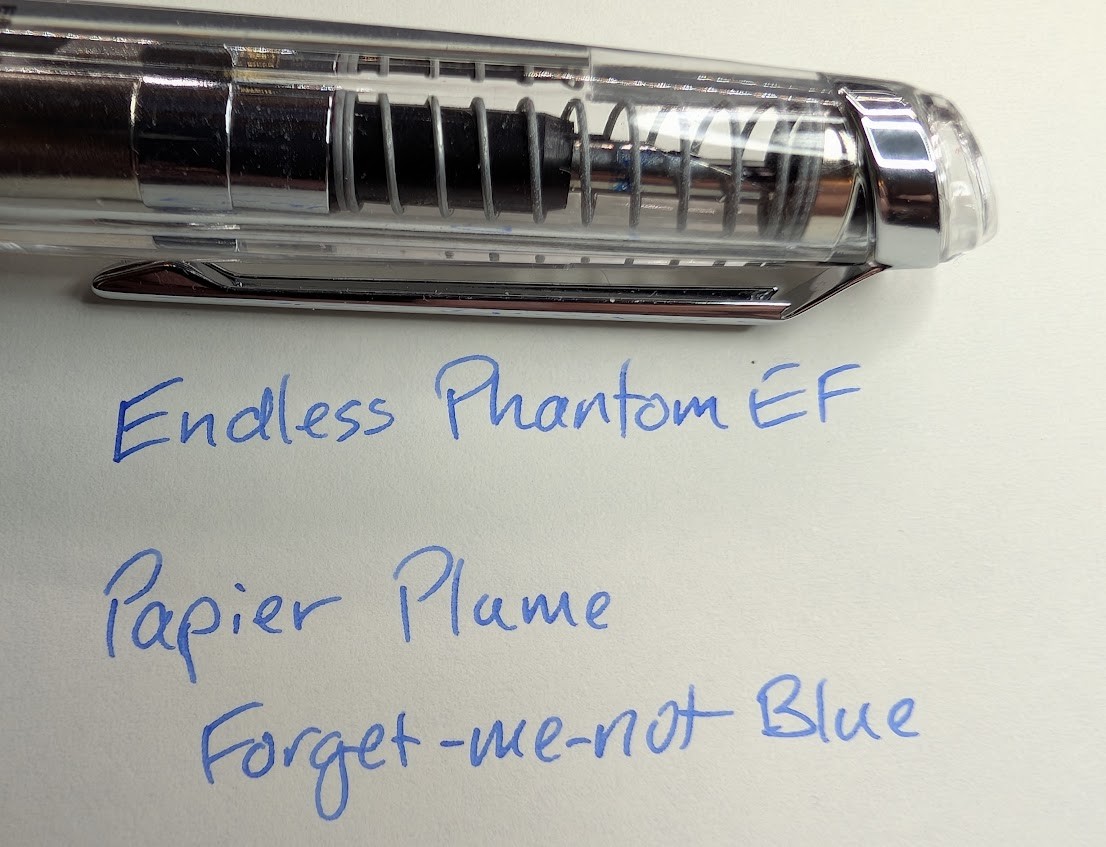 Photo of writing on paper with the tip of a capless fountain pen above it. The pen is transparent and the nib is visible through the body, surrounded by a spring. The writing reads "Endless Phantom EF / Papier Plume Forget-me-not Blue" in a bright medium blue ink.