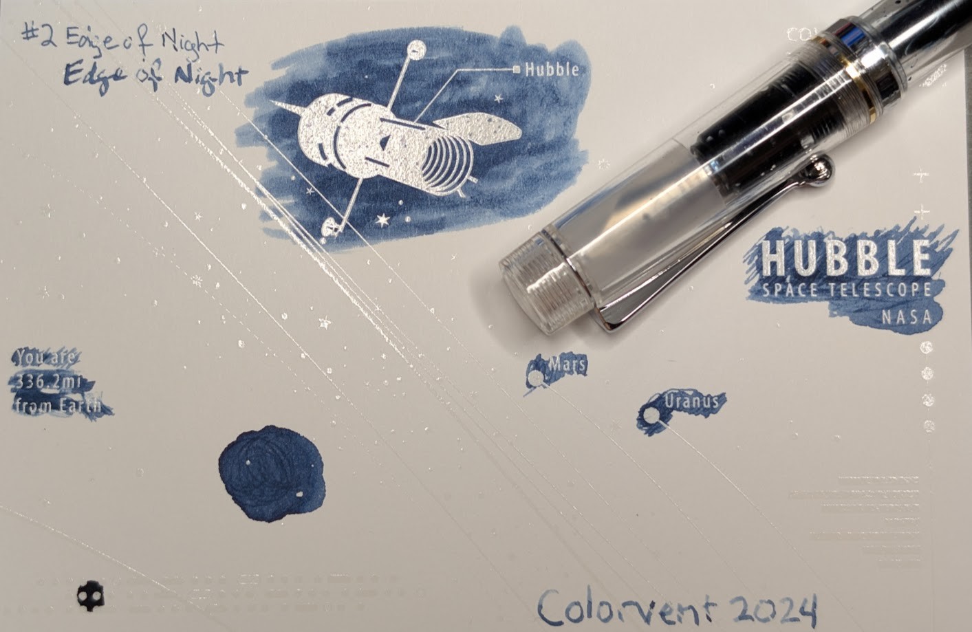 Photo of a "Colorverse Colorspace Size C" swatch card, type 2 which is based on the Hubble space telescope. The ink on the card is a blue-gray color that is light in shade, with some areas darker where ink has pooled. Upper left is text reading "#2 Edge of Night" written in an EF nib and then a stub nib. Just to the right side of the middle at the top is a large illustration of the Hubble space telescope in negative space among swabbed ink. In the middle right side is text "Hubble Space Telescope NASA" in negative space among swabbed ink. In the middle left is text reading "You are 336.2mi from Earth" in negative space among ink scribbles. In the middle lower left is a blob of swirled ink drawn by a glass dip pen. Near the center are "Mars" and "Uranus" with circles representing those planets, in more ink. Lower right is text "Colorvent 2024". Laying across the card is an Osprey Madison fountain pen, it is clear/transparent plastic with silver accents. Blue ink is visible in the section and body.