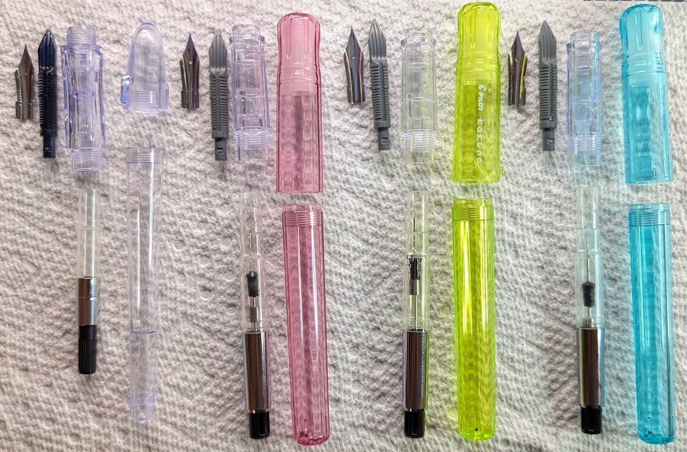 Photo of four disassembled fountain pens. Their caps, bodies, converters, sections, feeds, and nibs are all laid out next to each other in a row. From left to right they are: Pilot Penmanship, Pilot Kakuno Family "Girl" (Pink), Pilot Kakuno Family "Baby" (Yellow), and Pilot Kakuno Family "Boy" (Greenish/Teal)