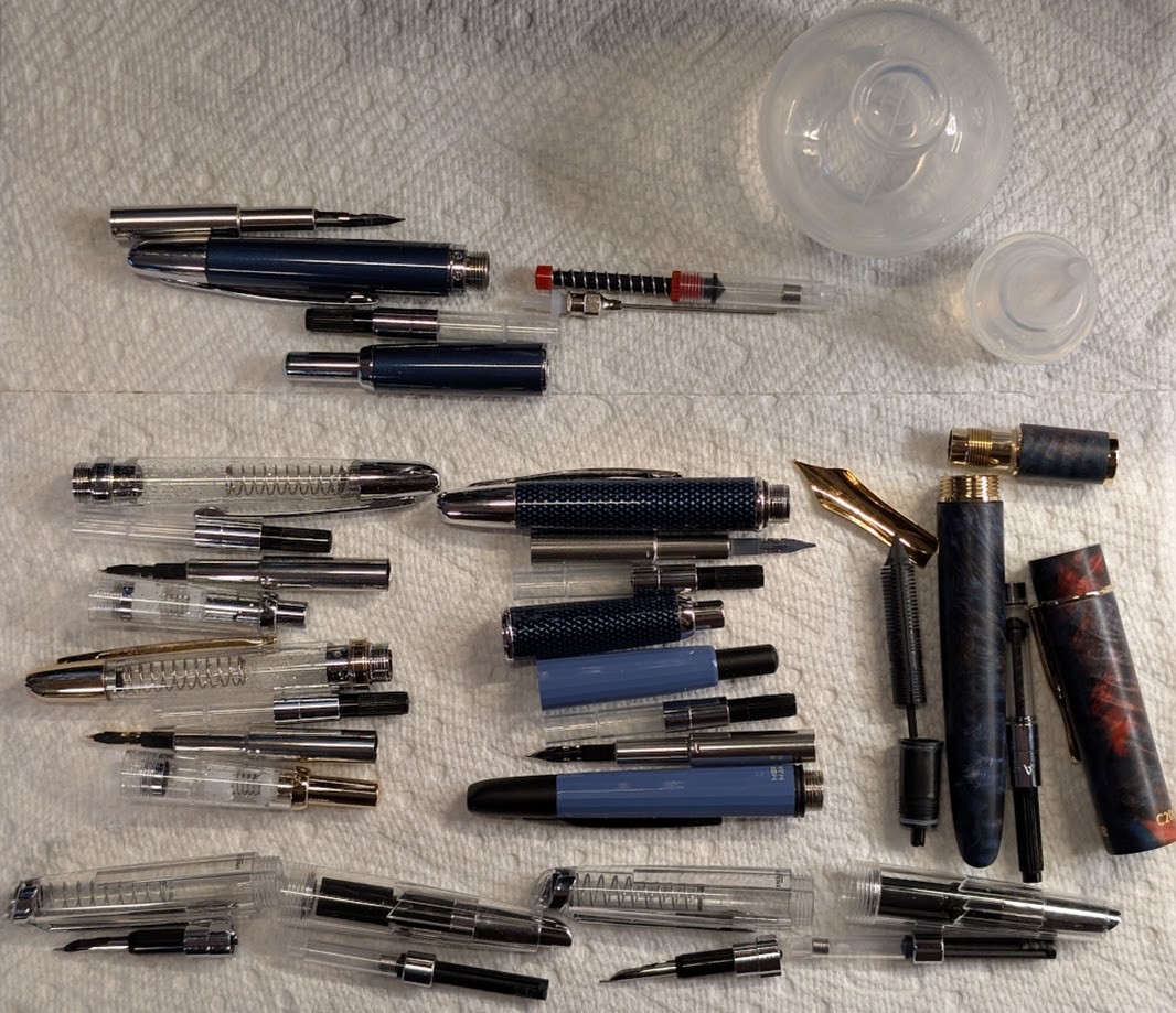 Photo of 8 disassembled fountain pens laid out on a paper towel. There is also a disassembled clear bulb syringe and a disassembled converter syringe. The pens are, in no particular order: Pilot VP, Asvine C2000, Majohn A1, Majohn A2, Jinhao 20 (x2), Endless Phantom (x2)