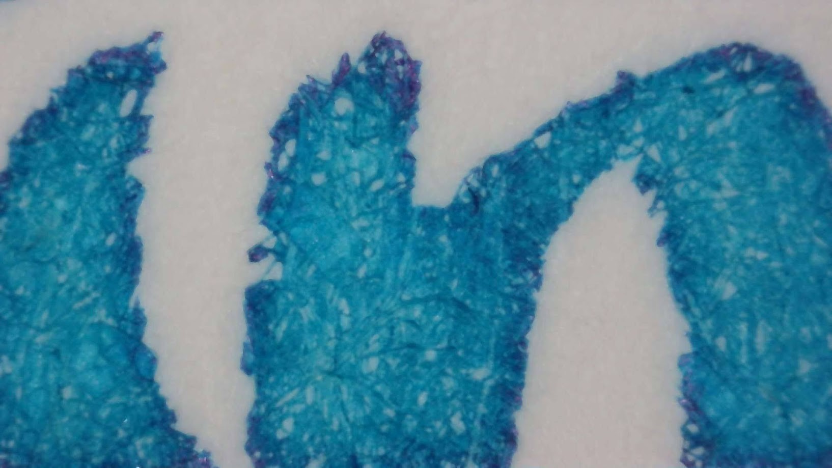 Photo of ink writing on paper under magnification. Paper fibers are visible. The ink is mostly medium blue-green but the edges are a darker shade and the very tips of the edges are reddish.