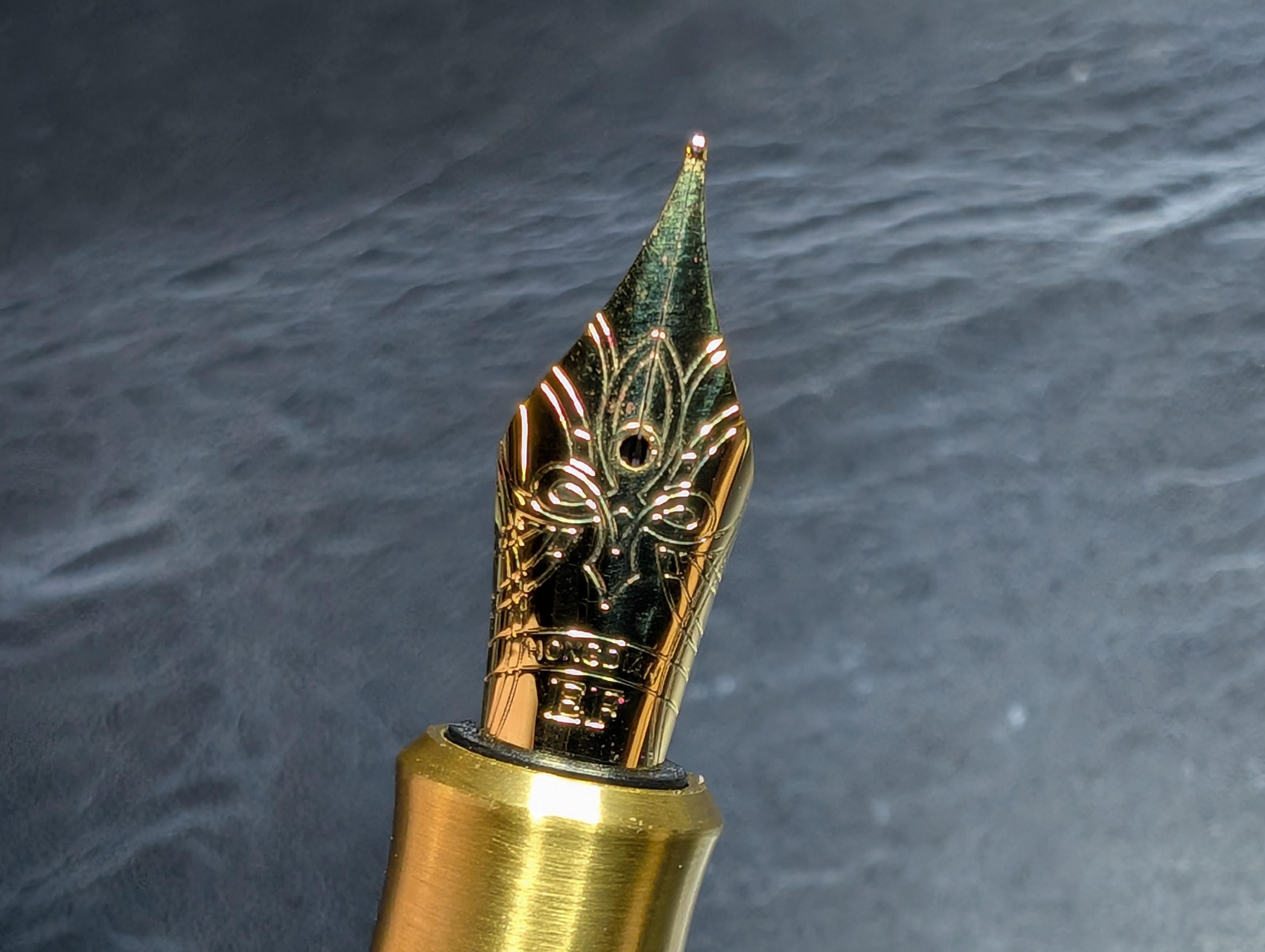 Photo of a fountain pen nib in a brass fountain pen. The nib is a similar brass tone to the pen itself. The nib has an elaborate Chinese knot design etched in it along with the words "Hongdian" and "EF"