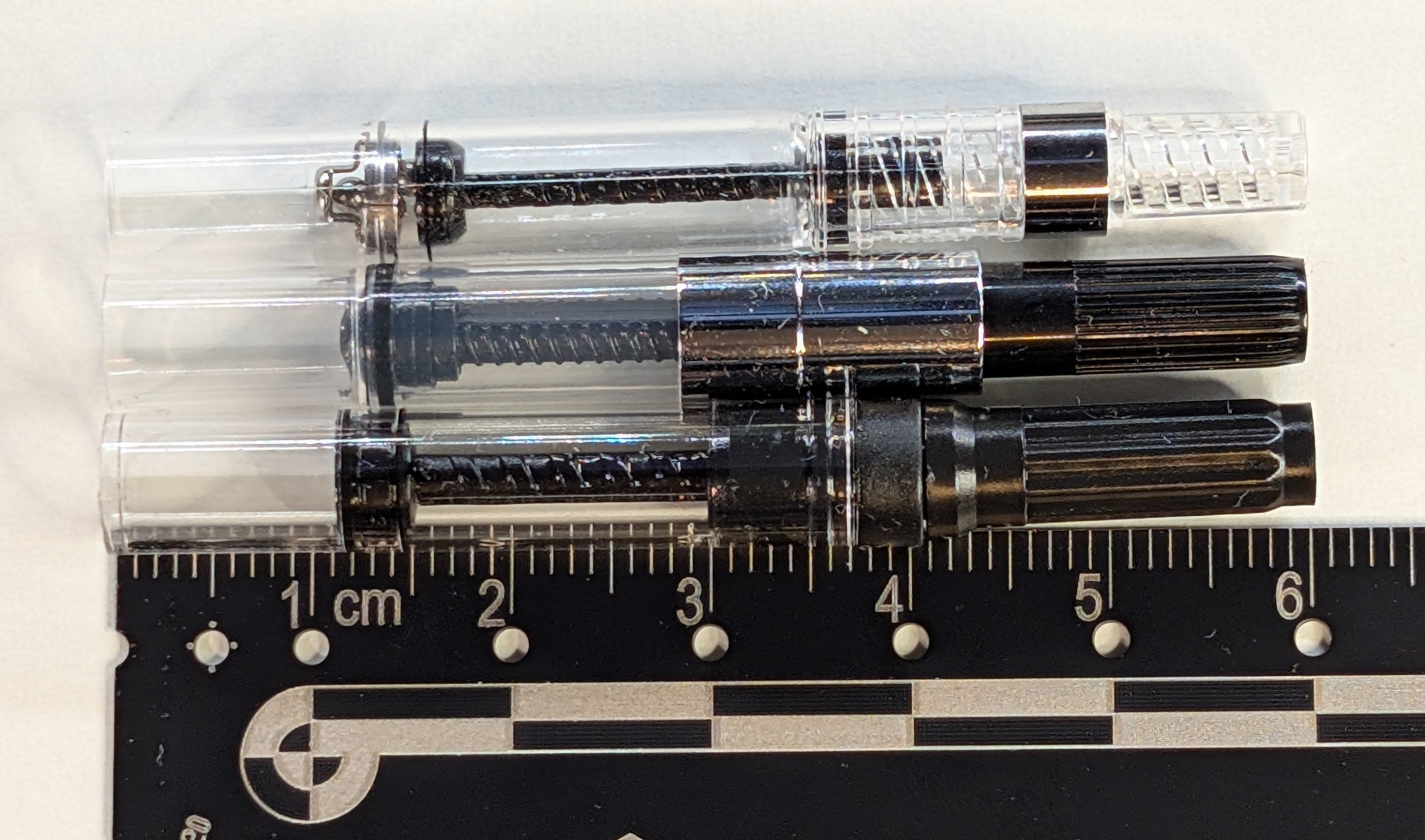 Photos of three fountain pen converters next to each other with their pistons extended, all next to a ruler. Top is a Pilot CON-40, the piston appears to be at about the 16mm mark on the ruler Next is a Majohn A1, the piston appears to be at about the 11mm mark Last is a Jinhao 10, the piston appears to be at about the 12mm mark
