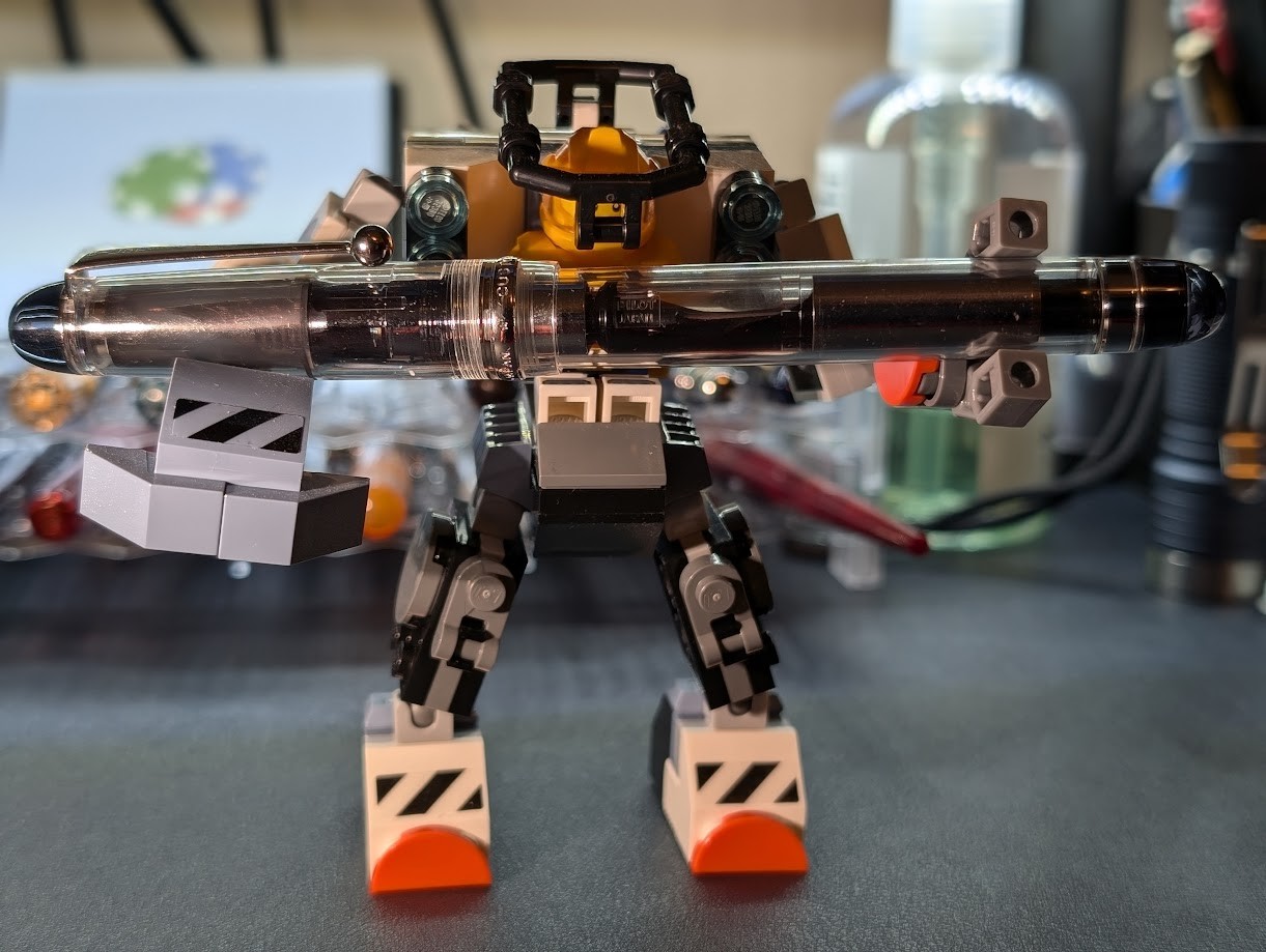 Photo of a fountain pen being held up by a mechanical suit made of Lego bricks. The fountain pen is a Pilot Custom 74 with a clear body and silver trim. It has rounded ends and a thin body. The Lego set holding up the pen is the "Space Construction Mech" #60428. It is a humanoid-shaped mech with a minifigure as the pilot. The pilot is wearing a yellow uniform and helmet, and is sitting with a "cage" over its head. One arm of the mech has pincers holding onto the pen. The other arm has a battering ram supporting the other end of the pen.