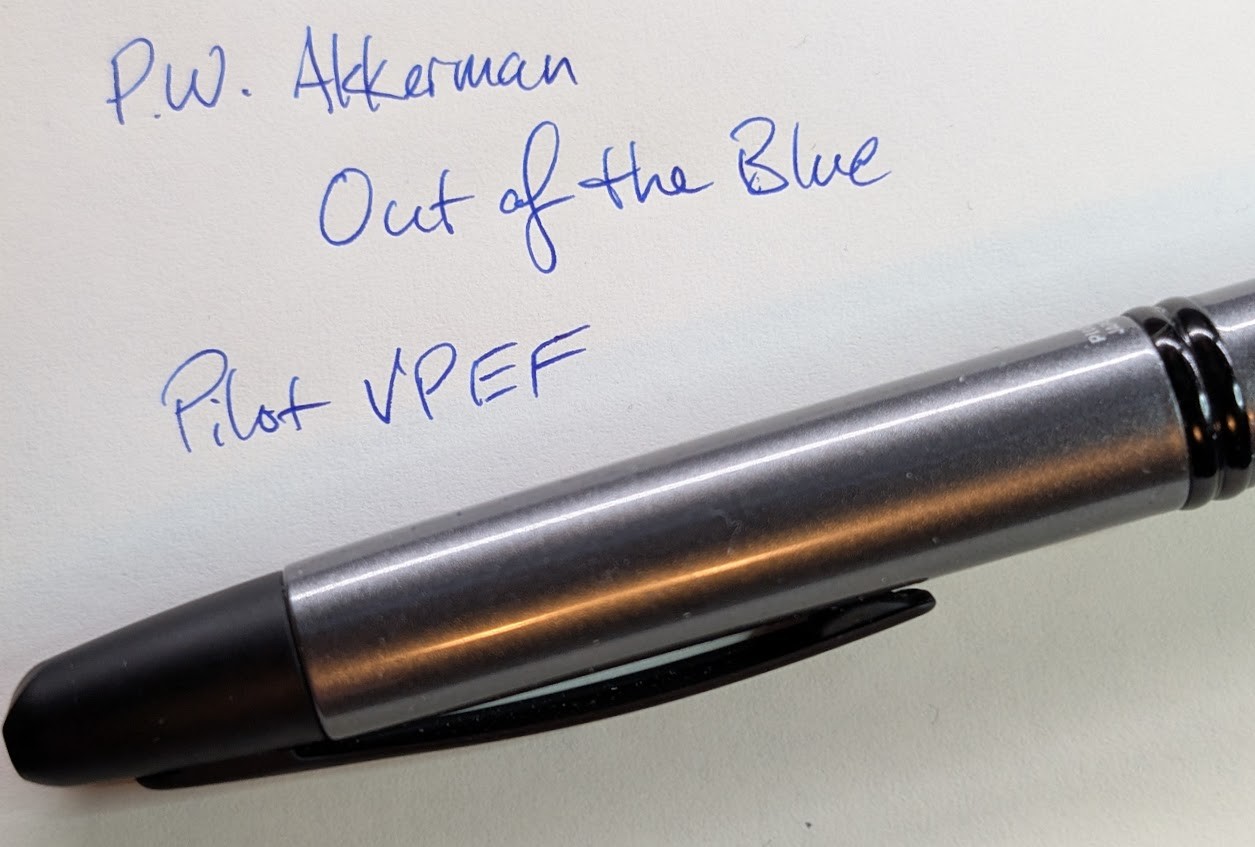 Photo of a dark gray metal capless fountain pen with matte black accents. It is sitting on a paper with writing in blue/purple ink which reads: "P.W. Akkerman / Out of the Blue / Pilot VP EF"