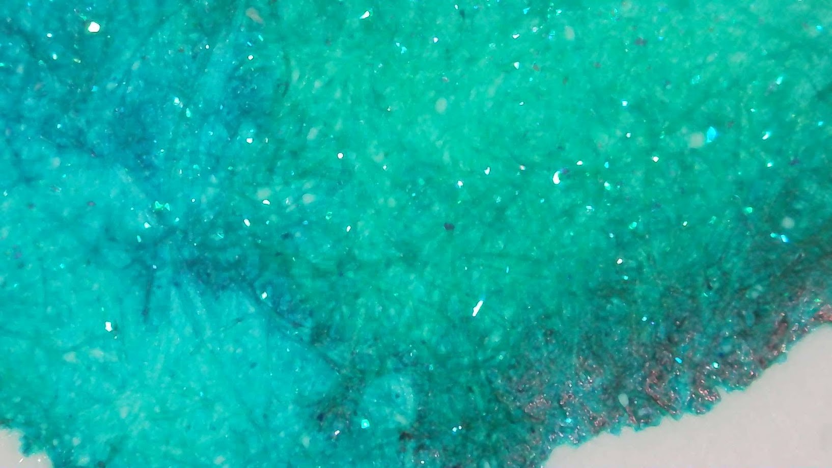 Photo of ink blob under magnification. Paper fibers are visible. The ink is a bright aqua/turquoise color. The left side is more blue, the right side is more green. There are cyan shimmer flakes occasionally throughout the blob.