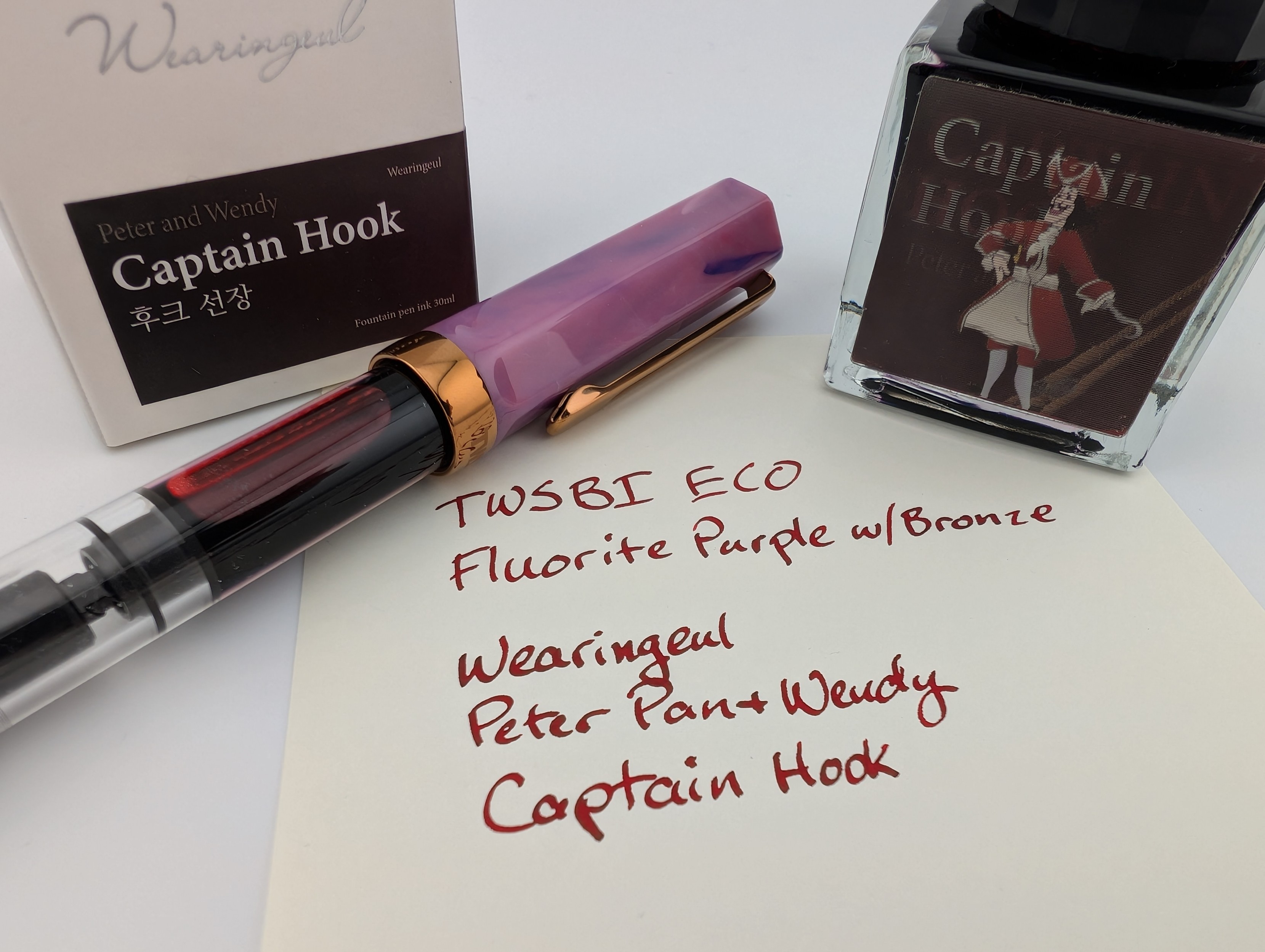 Photo of a TWSBI fountain pen (Fluorite Purple with Bronze) next to a fountain pen ink bottle and its box, and all of that is next to a writing sample. The ink is a dark red with some even darker shading. The writing sample reads: "TWSBI ECO / Fluorite Purple with Bronze / Wearingeul / Peter Pan and Wendy / Captain Hook." The ink bottle has a lenticular label and shows both "Captain Hook" in text and an image of the captain.