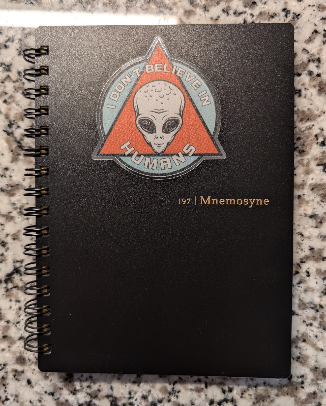 Photo of a black spiral-bound notebook on a desk. It is labeled "197 | Mnemosyne". There is a sticker on top that has the face of an alien with text surrounding it reading: "I don't believe in humans"