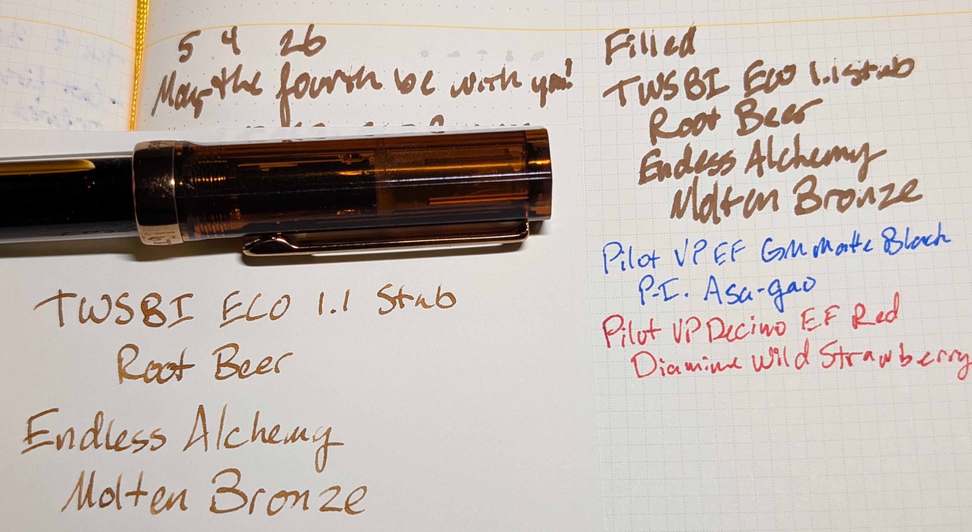 Photo of a fountain pen resting on an open notebook and a separate sheet of paper. The fountain pen is a TWSBI ECO in the "Root Beer" colorway which is has a transparent brown cap and bronze trim and clip. The sheet of paper has a writing sample reading "TWSBI ECO 1.1 Stub / Root Beer / Endless Alchemy Molten Bronze" in brown ink. The lines are easy to read, clean, and evenly colored with some variation in width. The notebook has some writing with the same ink reading "5 . 4 . 26 / May the fourth be with you!" and "Filled / TWSBI ECO 1.1 Stub / Root Beer / Endless Alchemy Molten Bronze". The lines are much wider, fuzzier, and hard to read because they run together due to feathering. Under those are two unrelated writing samples for a Pilot VP in blurple Asa-gao ink and a Pilot VP Decimo in red Diamine Wild Strawberry ink.