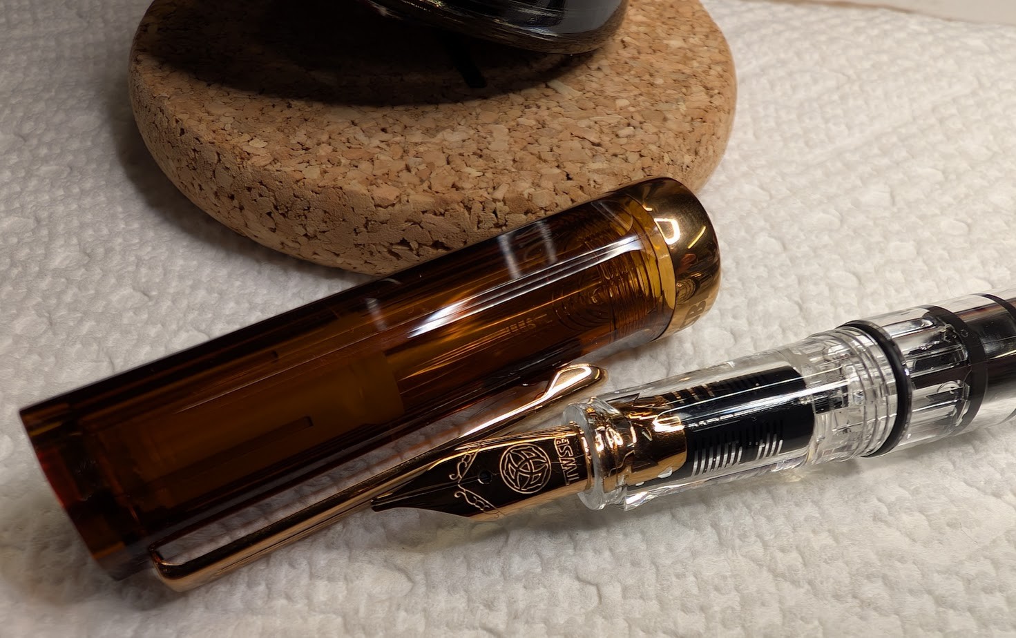 Photo of a fountain pen on a paper towel next to an ink bottle. The fountain pen is a TWSBI ECO in the "Root Beer" colorway which is has a transparent brown cap and bronze trim and clip. The cap is removed and next to the pen. The nib is also bronze. The section and body of the pen are clear. The ink bottle is barely visible, but is an Endless Alchemy bottle which is flask-shaped and resting on a cork pad.