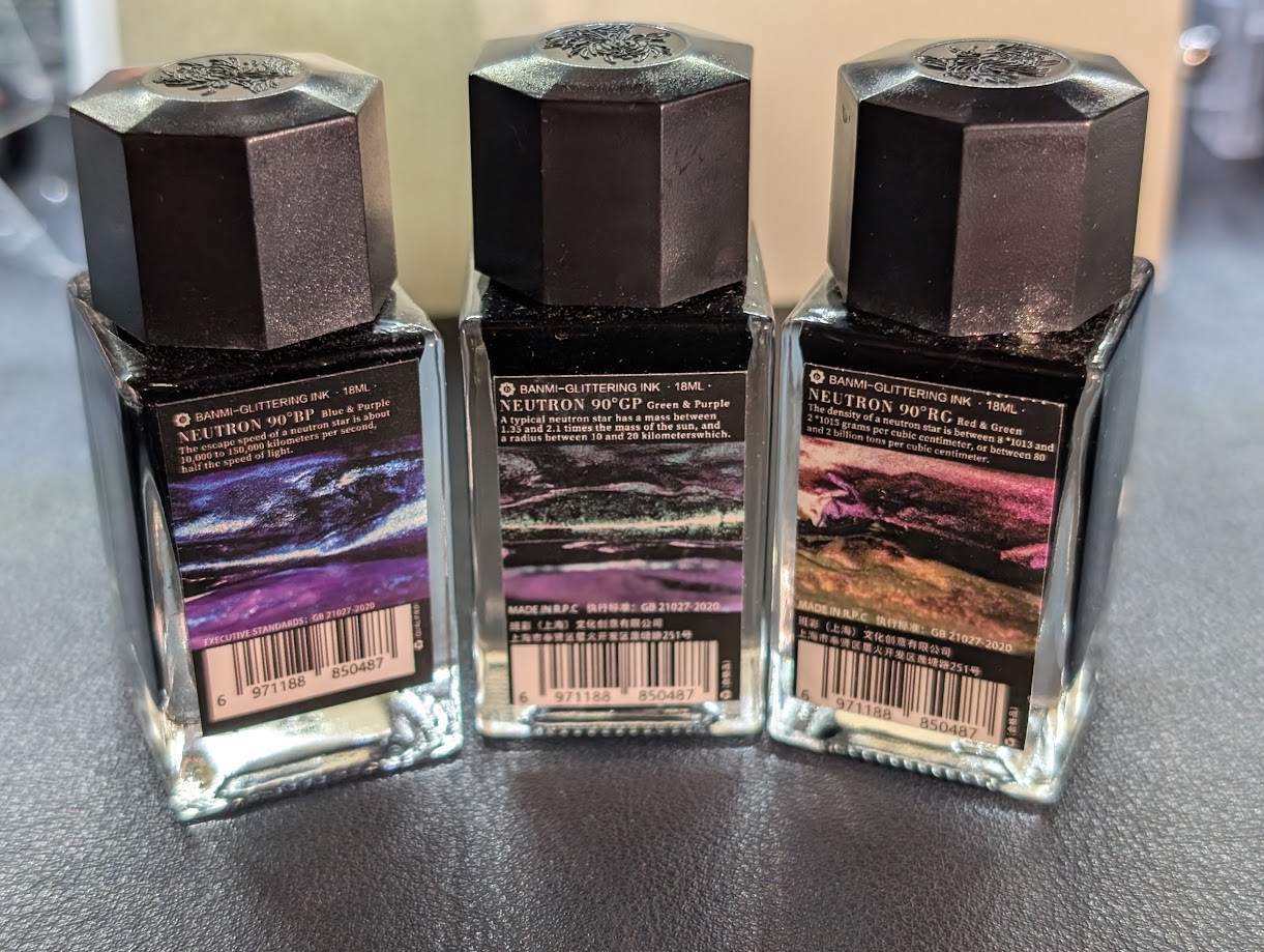 Photo of three 18ml bottles of fountain pen inks. Labels say: Banmi Glittering Ink 18ml, Neutron 90°. Each bottle has a different name and text. From left to right they are BP (Blue & Purple), GP (Green & Purple), RG (Red & Green).