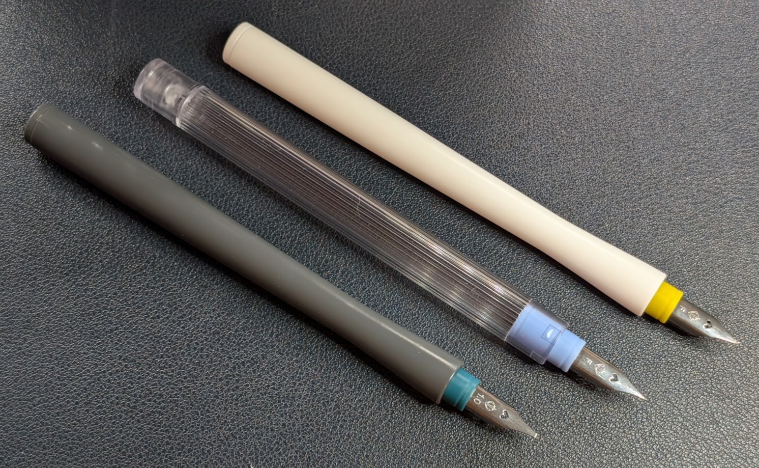 Photo of three dip pens on a table. From left to right they are: - Gray body, green 1.0 stub nib labeled "1.0". - Clear body, light blue Fine nib labeled "F". - White body, yellow Fude nib labeled "F".