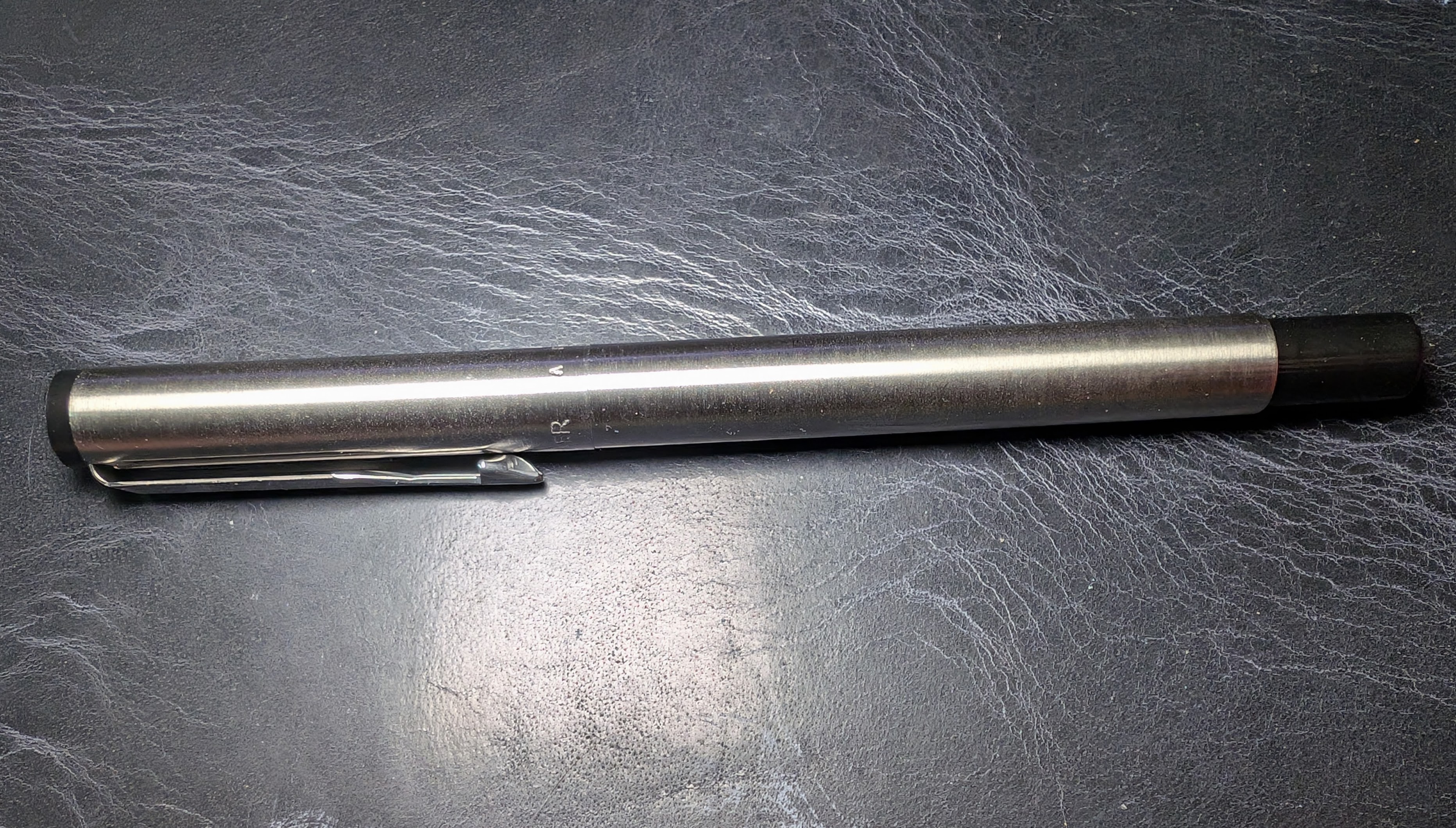 Photo of a stainless steel fountain pen on a dark blue leather surface. The pen is very narrow and has perfectly straight sides. The finial and tail are black plastic.