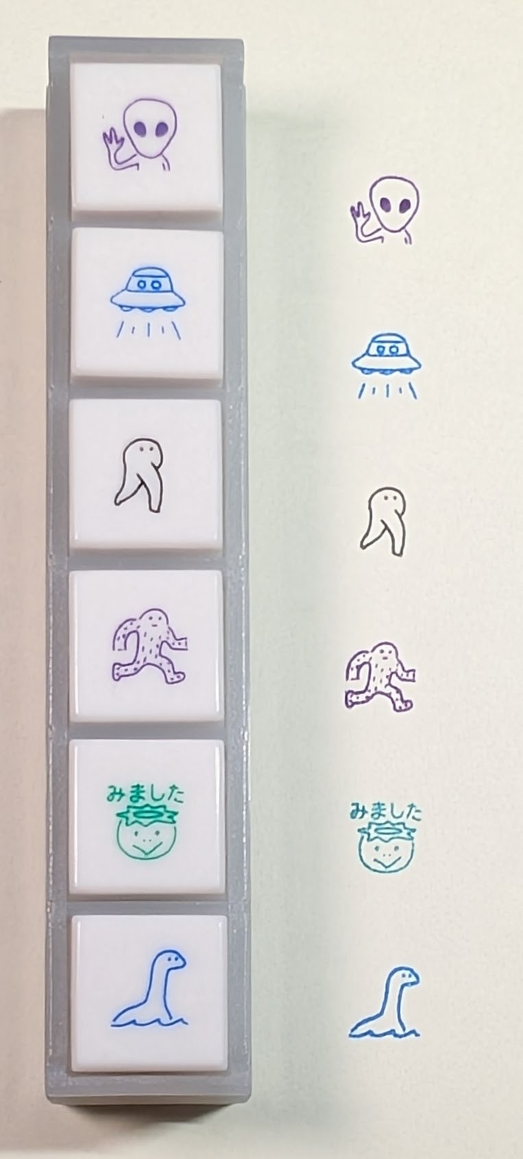 A row of six linked cryptid stamps next to their stamp marks on paper. The marks match the icon on the stamp. From top to bottom they are: - A purple "gray" type alien with one hand up waving - A blue UFO - A gray Nightwalker - A purple Bigfoot - A green Kappa with "I saw it" written over it in Japanese - A blue Nessie
