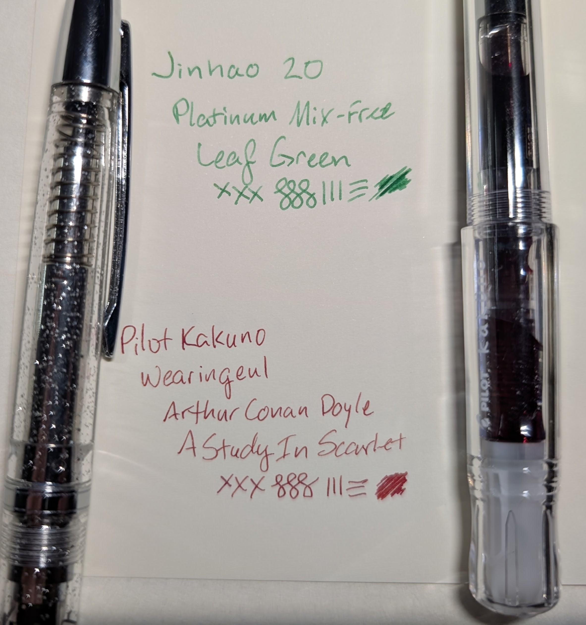 Photo of two pens and writing samples on a piece of paper. Top is in medium green ink reading "Jinhao 20 / Platinum Mix-Free / Leaf Green" followed by some lines and scribbles. To the left is a Jinhao 20 fountain pen which is transparent with silver accents and some shimmer in the plastic. Bottom is in dark red ink reading "Pilot Kakuno / Wearingeul / Arthur Conan Doyle / A Study in Scarlet" followed by some lines and scribbles. To the right is a Pilot Kakuno fountain pen with a transparent body and cap.