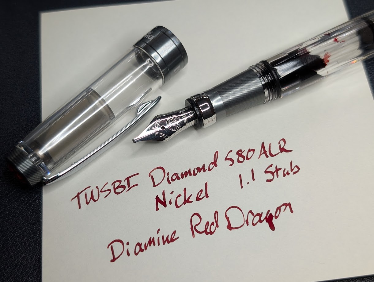 Photo of a fountain pen and writing sample. The fountain pen is a TWSBI Diamond 580ALR in Nickel, with silver trim. The finial, bottom rim of the cap, and section are all gray. The cap and body of the pen are clear. Red ink is visible inside. The writing sample is in dark red ink and reads "TWSBI Diamond 580ALR Nickel 1.1 Stub" and "Diamine Red Dragon"