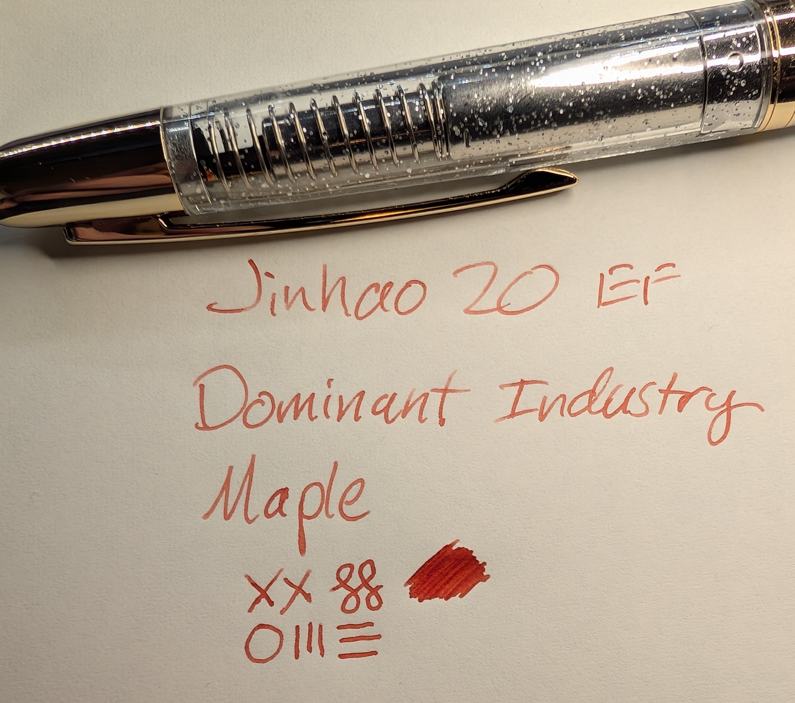 Photo of a fountain pen and writing on paper. The writing reads "Jinhao 20 EF / Dominant Industry / Maple" followed by "XX", two curlicues, a circle, three vertical lines, three horizontal lines, and a scribbled blob of ink. The ink is dark orange that leans a little red. The fountain pen is a Jinhao 20 that has a transparent body with a little shimmer in the plastic, and gold trim. The spring and nib are visible through the body.