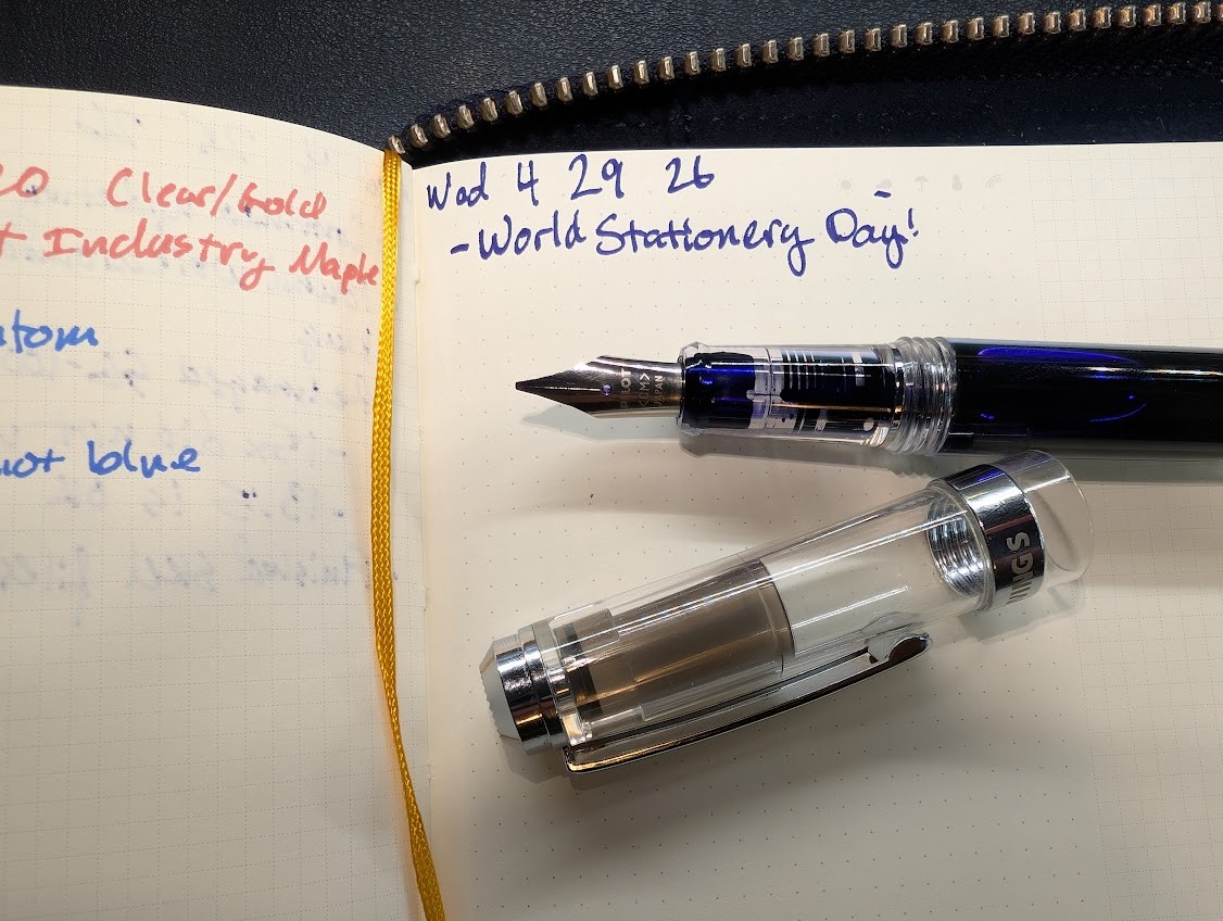 Photo of an uncapped fountain pen resting on a notebook which is in a leather zipper folio. The page reads "Wed 4 29 26 - World Stationery Day!" in purple ink (Diamine Bilberry) The pen is a WingSung 698 but with a Pilot CM nib. On the opposing page are two bits of writing which are partially visible, the wording is not important but the lines are very thick and with rough edges, and the lines are thicker then the writing on the text quoted above.