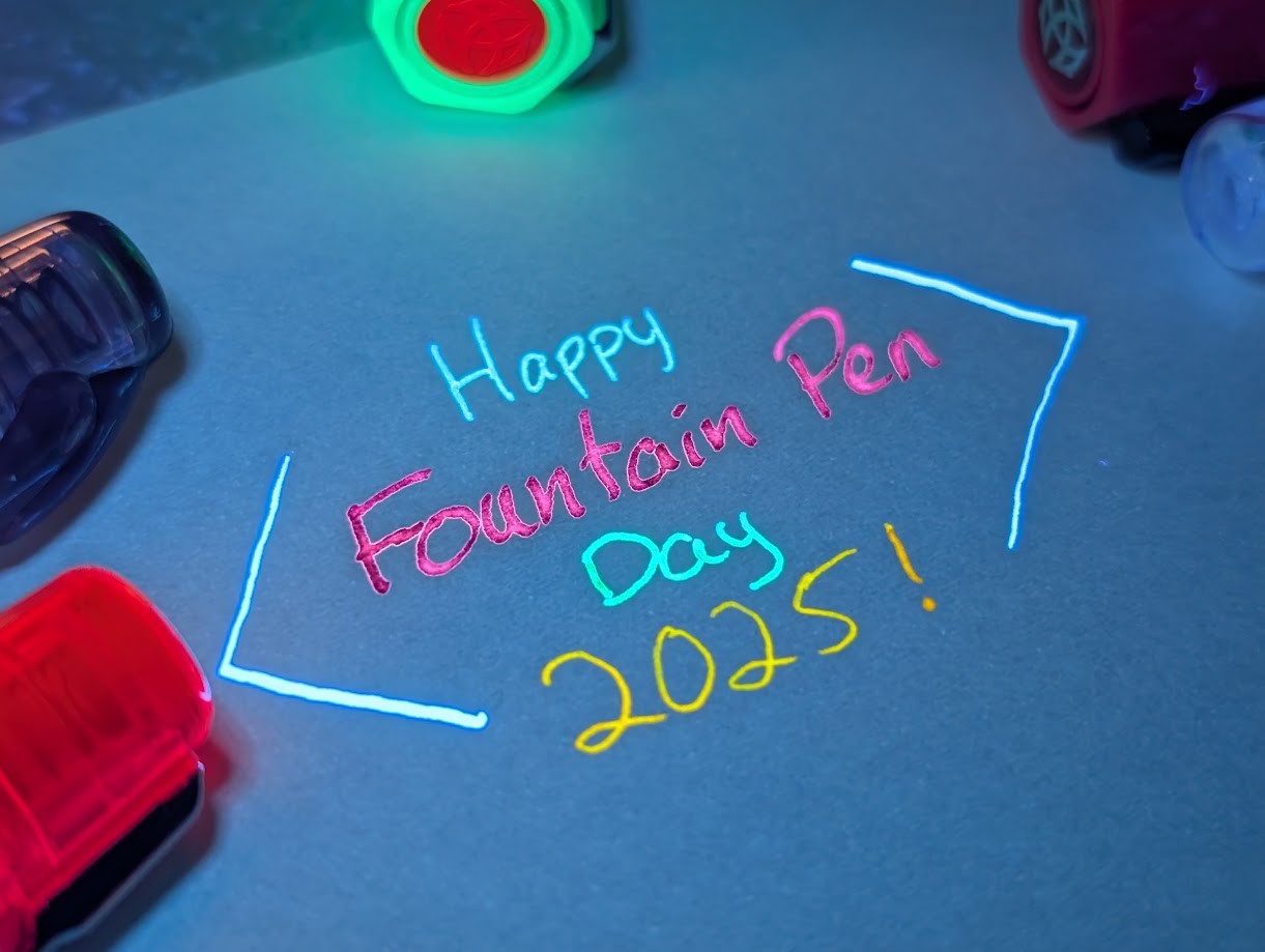 Photo of writing on paper at an oblique angle surrounded by the caps of several pens all under UV light. It reads "Happy Fountain Pen Day 2025!" with angled brackets around it. "Happy" is written in brightly glowing blue ink. "Fountain Pen" is written in brightly glowing pink ink "Day" is written in brightly glowing green ink "2025!" is written in brightly glowing orange ink The angle brackets are drawn with brightly glowing light blue ink The pens are, clockwise from lower left, a glowing orange Prefounte, a Preppy, a brightly glowing green TWSBI ECO with a red finial, a fuchsia TWSBI ECO, and another Preppy.