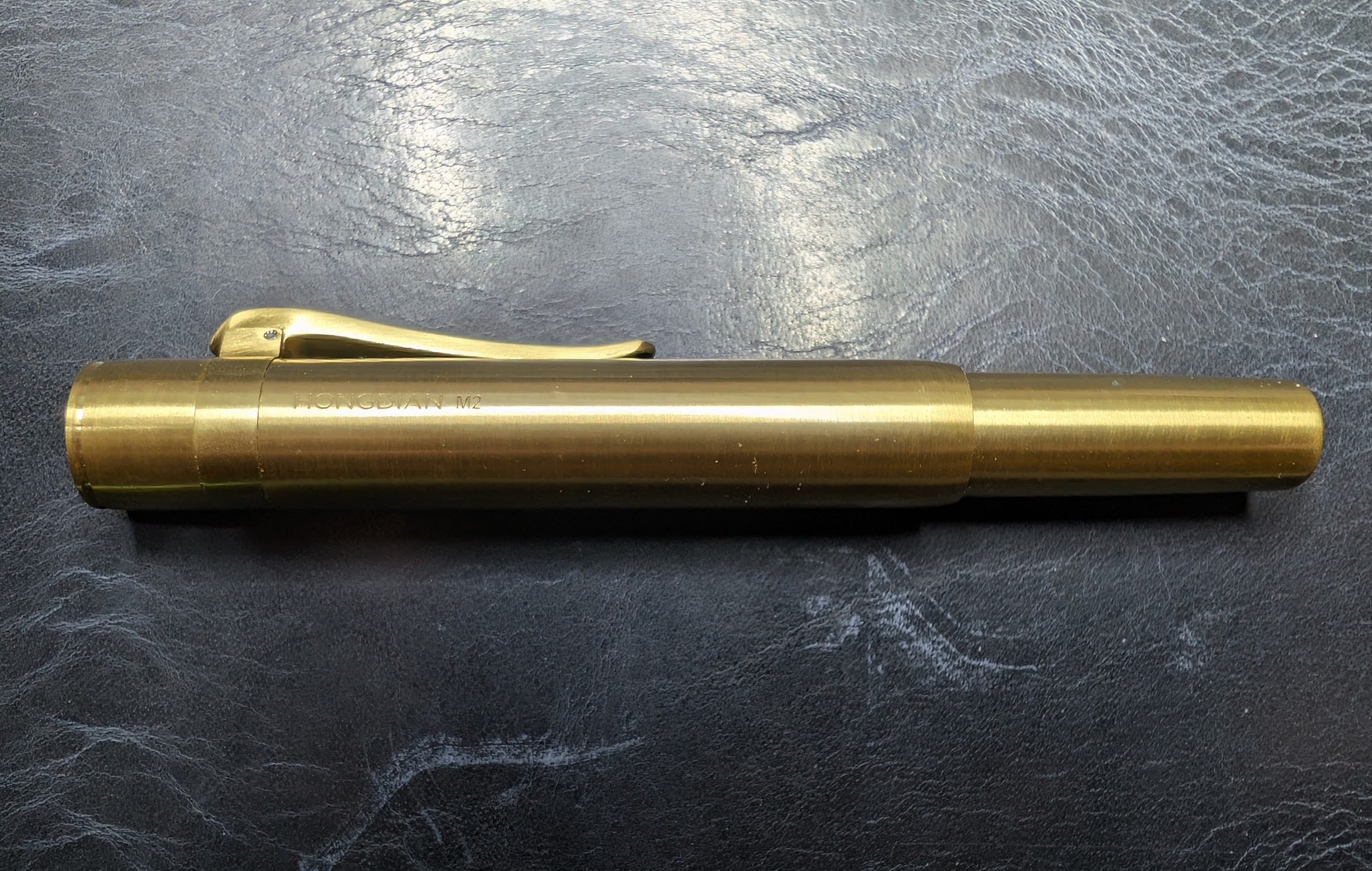 Photo of a brass fountain pen on a dark blue leather surface. It's shorter than a typical pen and its cap covers a large portion of its length. Laser etched on the side is some text reading "Hongdian M2"