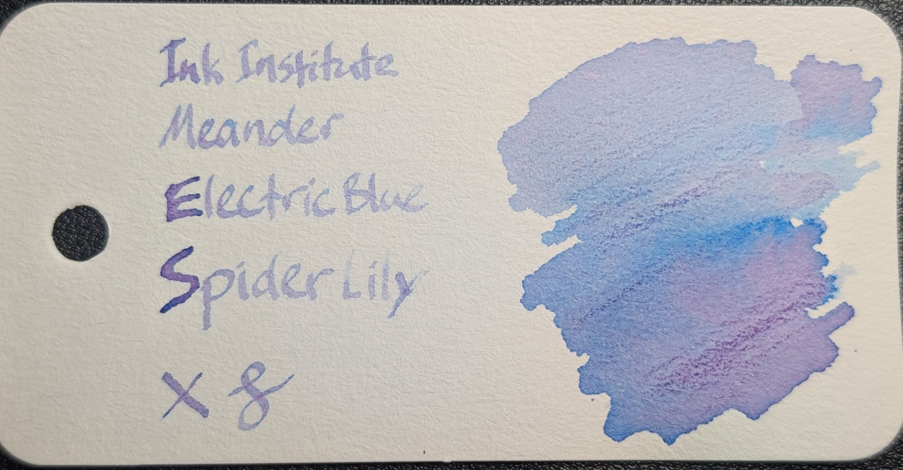 Photo of a col-o-ring swatch card. The ink on the card is an ink that appears both light blue and pink in different places. On the right is a large ink blob that is light blue in places with less ink, and in places with more ink it's pink with dark blue edges. On the left is text reading "Ink Institute Meander Electric Blue Spider Lily" with an X and a curlicue underneath. The text is light, but legible. Some letters are darker and in those letters the edges are darker blue and the center is pink.