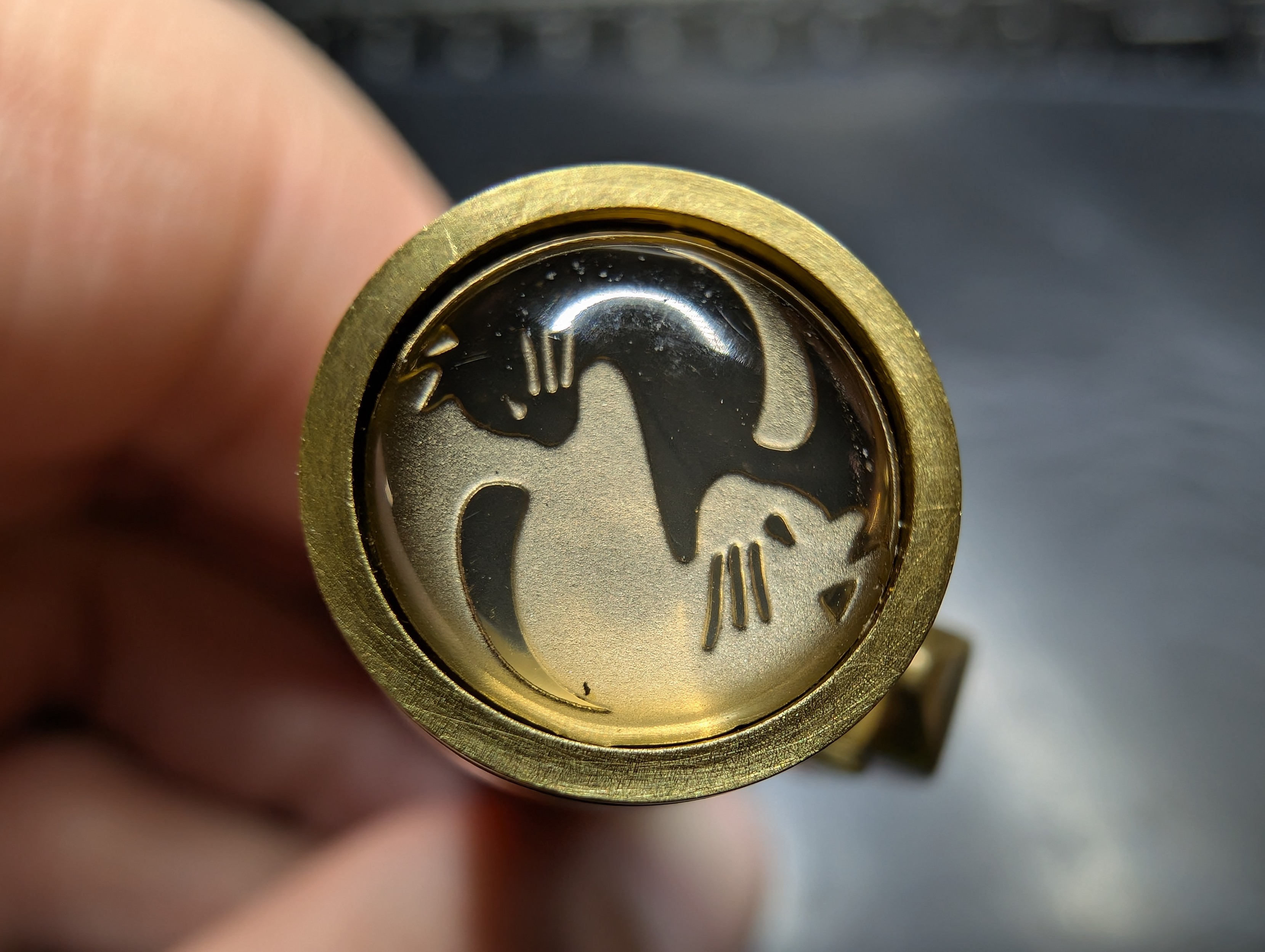 Photo of a brass fountain pen finial. It contains a design with a dark and a light cat curled together in a vaguely yin-yang style pattern. The brass gives the cats a bit of a yellow cast. The finial design is covered in a clear glossy dome.