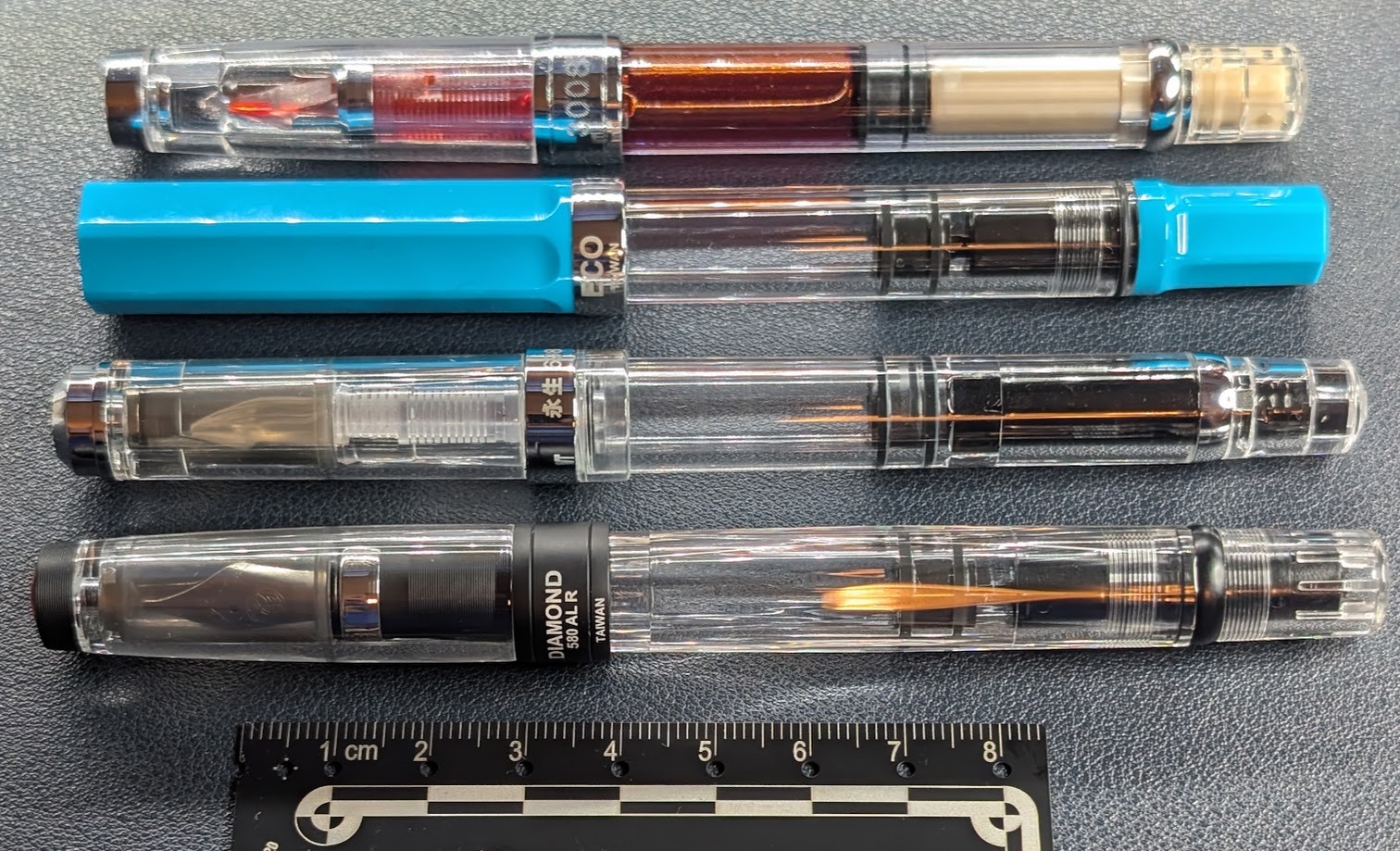 Photo of four fountain pens: From top to bottom: WingSung 3008 -- Clear with silver accents. Clear smooth round cap. Clear smooth round body, filled with red ink. Piston plunger is tan. Piston knob is clear. TWSBI ECO -- Cerulean Blue with Silver accents. Flat faceted opaque cap is cerulean blue. Clear smooth round body, no ink inside. Piston plunger is black. Piston knob is opaque cerulean blue. WingSung 698 -- Clear with silver accents. Clear smooth round cap. Clear smooth round body, no ink inside. Piston plunger is black. Piston knob is clear. TWSBI Diamond 580 ALR -- Clear with black and silver accents. Clear smooth round cap. Clear diamond faceted body, no ink inside. Piston plunger is black. Piston knob is clear. Beneath the photo is a centimeter ruler with a photogrammetric scale.