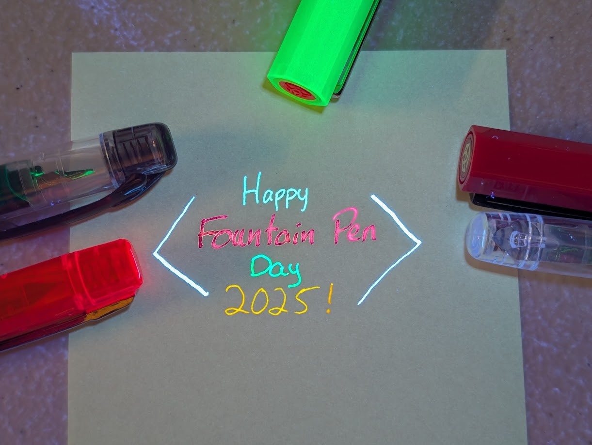 Photo of writing on paper surrounded by the caps of several pens all under UV light. It reads "Happy Fountain Pen Day 2025!" with angled brackets around it. "Happy" is written in brightly glowing blue ink. "Fountain Pen" is written in brightly glowing pink ink "Day" is written in brightly glowing green ink "2025!" is written in brightly glowing orange ink The angle brackets are drawn with brightly glowing light blue ink The pens are, clockwise from lower left, a glowing orange Prefounte, a Preppy, a brightly glowing green TWSBI ECO with a red finial, a fuchsia TWSBI ECO, and another Preppy.
