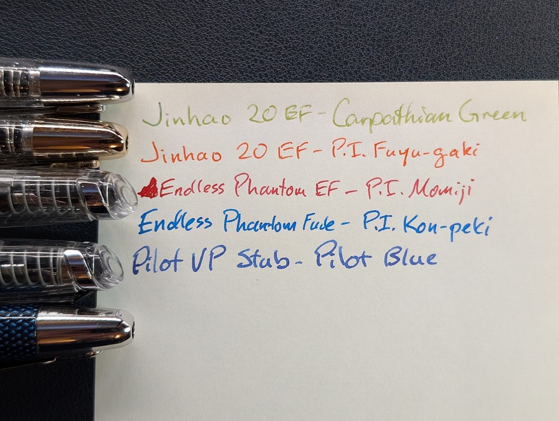 Photo of writing samples on paper with the tips of corresponding pens visible to the left. The samples are: - "Jinhao 20 EF - Carpathian Green" in very light olive-ish green. - "Jinhao 20 EF - P.I. Fuyu-gaki" in orange ink - "Endless Phantom EF - P.I. Momiji" in red ink - "Endless Phantom Fude - P.I. Kon-peki" in blue ink - "Pilot VP Stub - Pilot Blue" in dark blue ink
