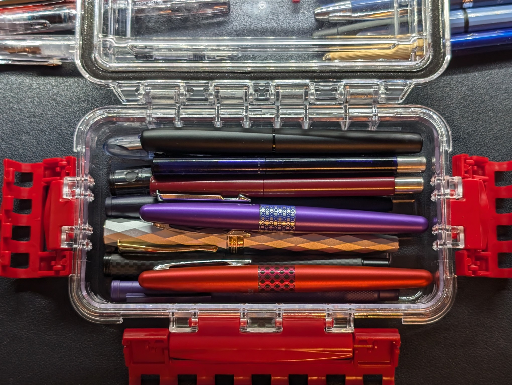 Photo of a clear Plano case with red latches. The case is open. Inside the case are about 10 fountain pens of various makes, models, colors, and styles.
