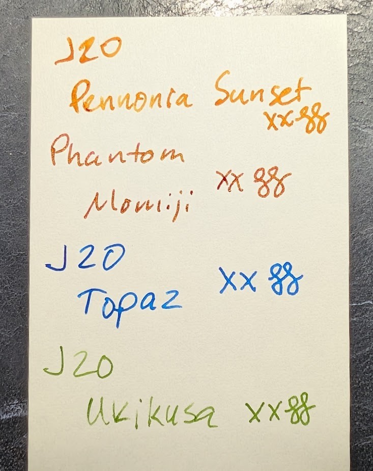 Four writing samples on a thin strip of Midori MD paper. Each is followed by "XX" and two curlicues. - "J20 Pennonia Sunset" in orange ink - "Phantom Momiji" in red ink with gold sheen, which is partially visible. - "J20 Topaz" in blue ink - "J20 Ukikusa" in olive green ink