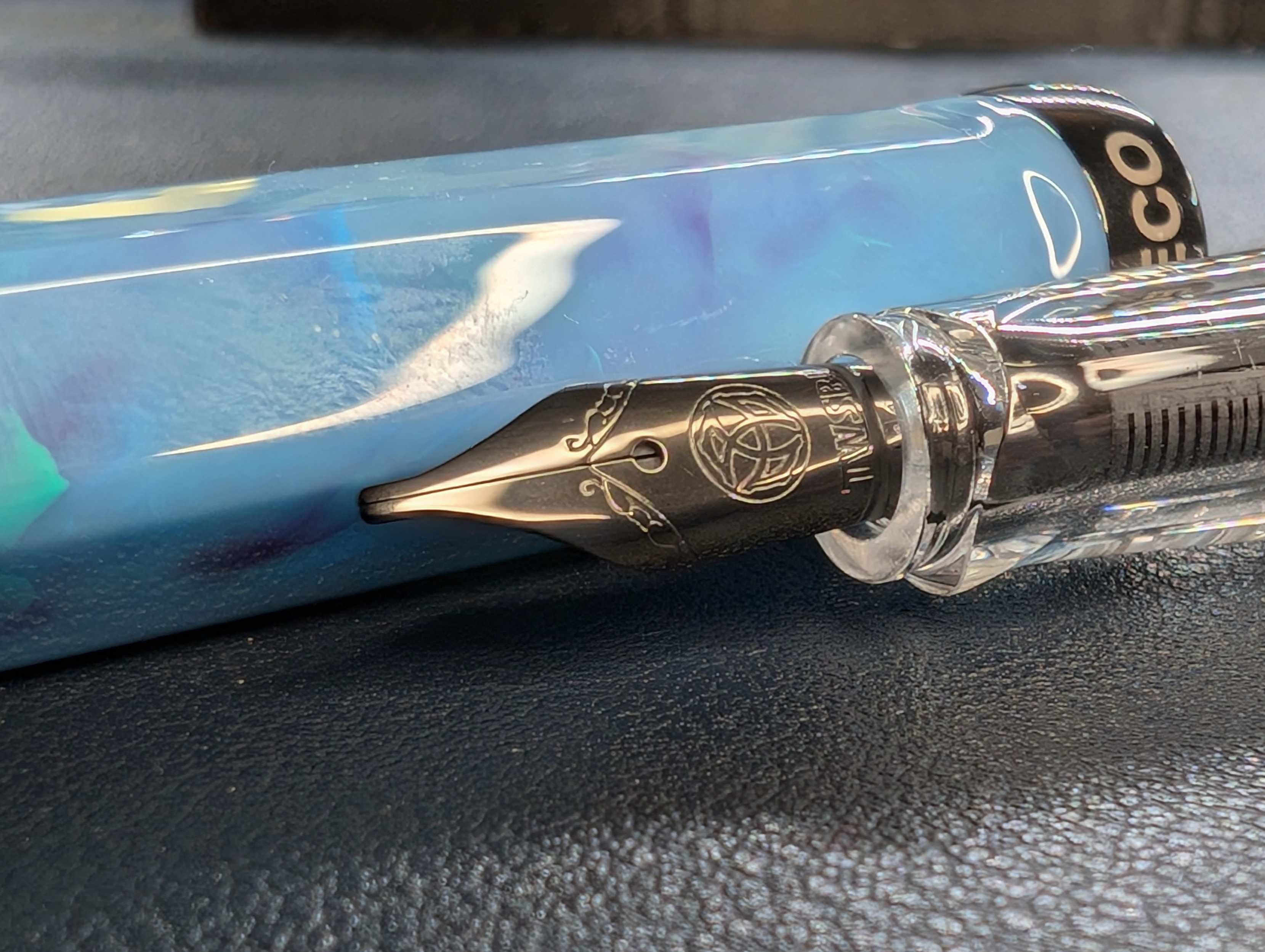 Photo of an uncapped fountain pen with the cap laying next to the pen. The nib is dark metal, almost black. The pen cap is a swirled mix of blue with streaks of purple and green.