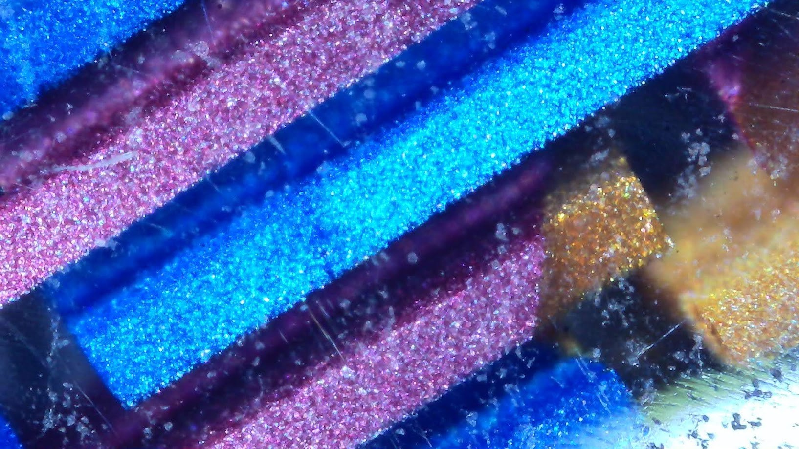 Photo of a fountain pen under a microscope. There are rectangular bars of color with a square cross-section going from the lower left to upper right. They are mostly blue and purple, with one partially gold. They appear to be suspended in clear resin. Some surface scratches and pits are visible.