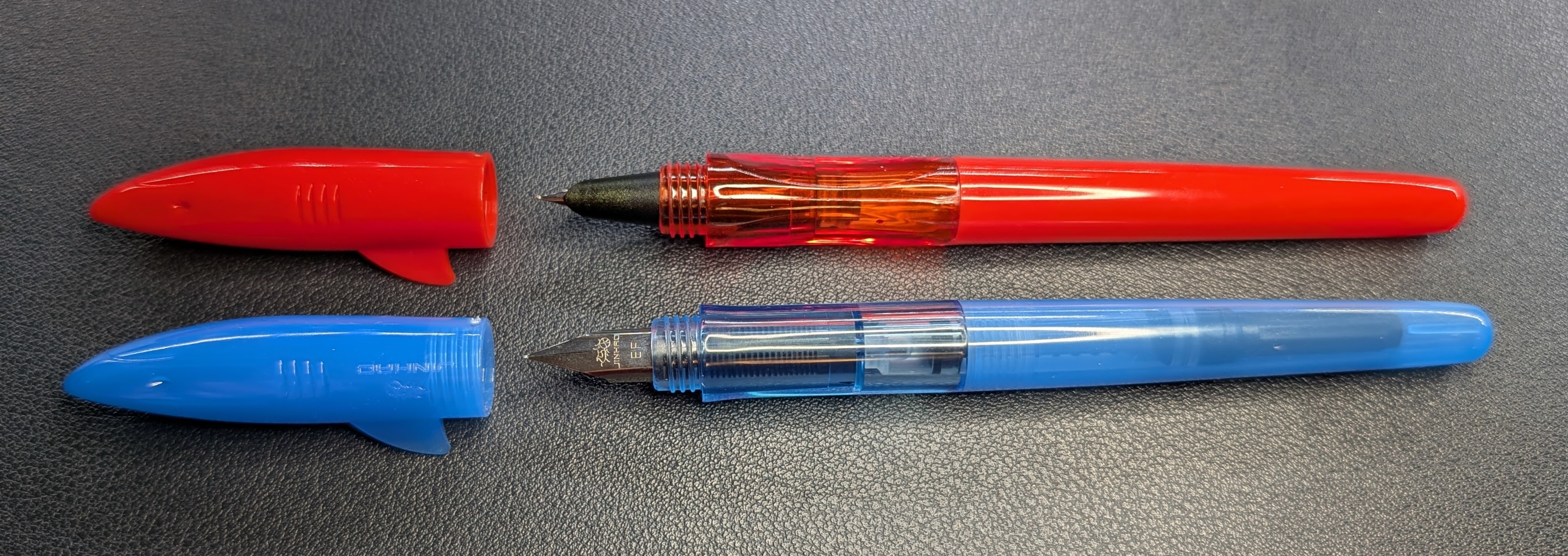 Photo of two Jinhao Shark fountain pens on a desk. The top pen is an older model, opaque red, and has a hooded nib and indents on the section where fingers rest when gripping. The bottom pen is new, transparent blue, and has an exposed nib and smooth, rounded section.